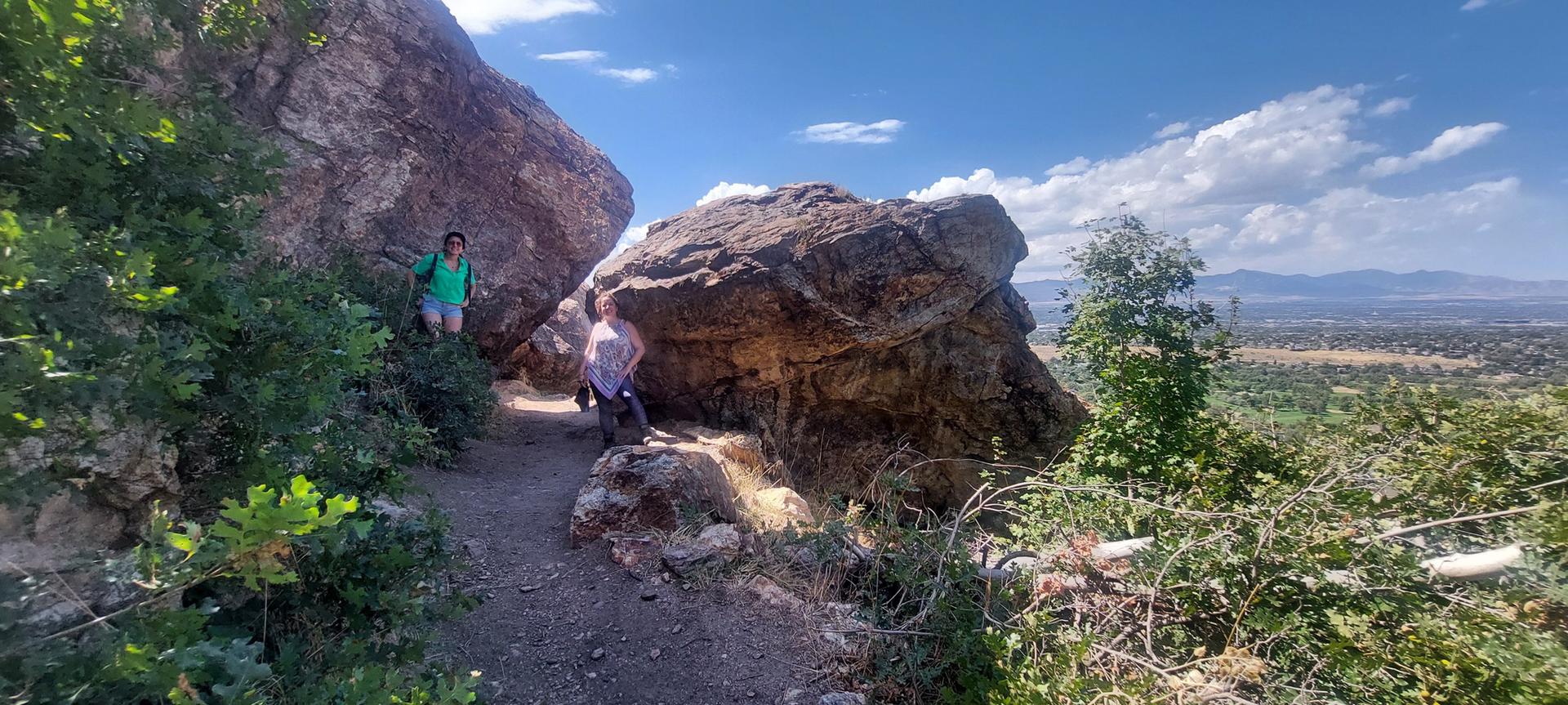 Two hikers navigate a rocky trail between large boulders on a sunny day.