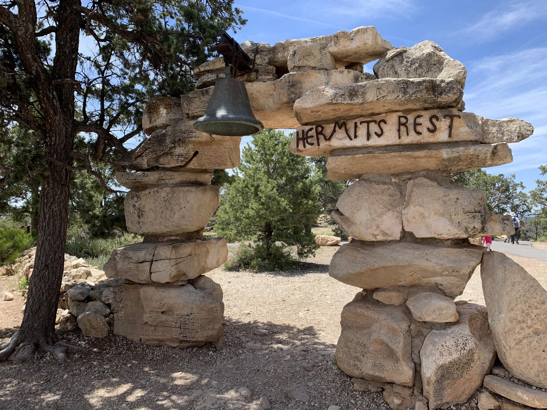 Stone archway with a bell and carved text, surrounded by trees.