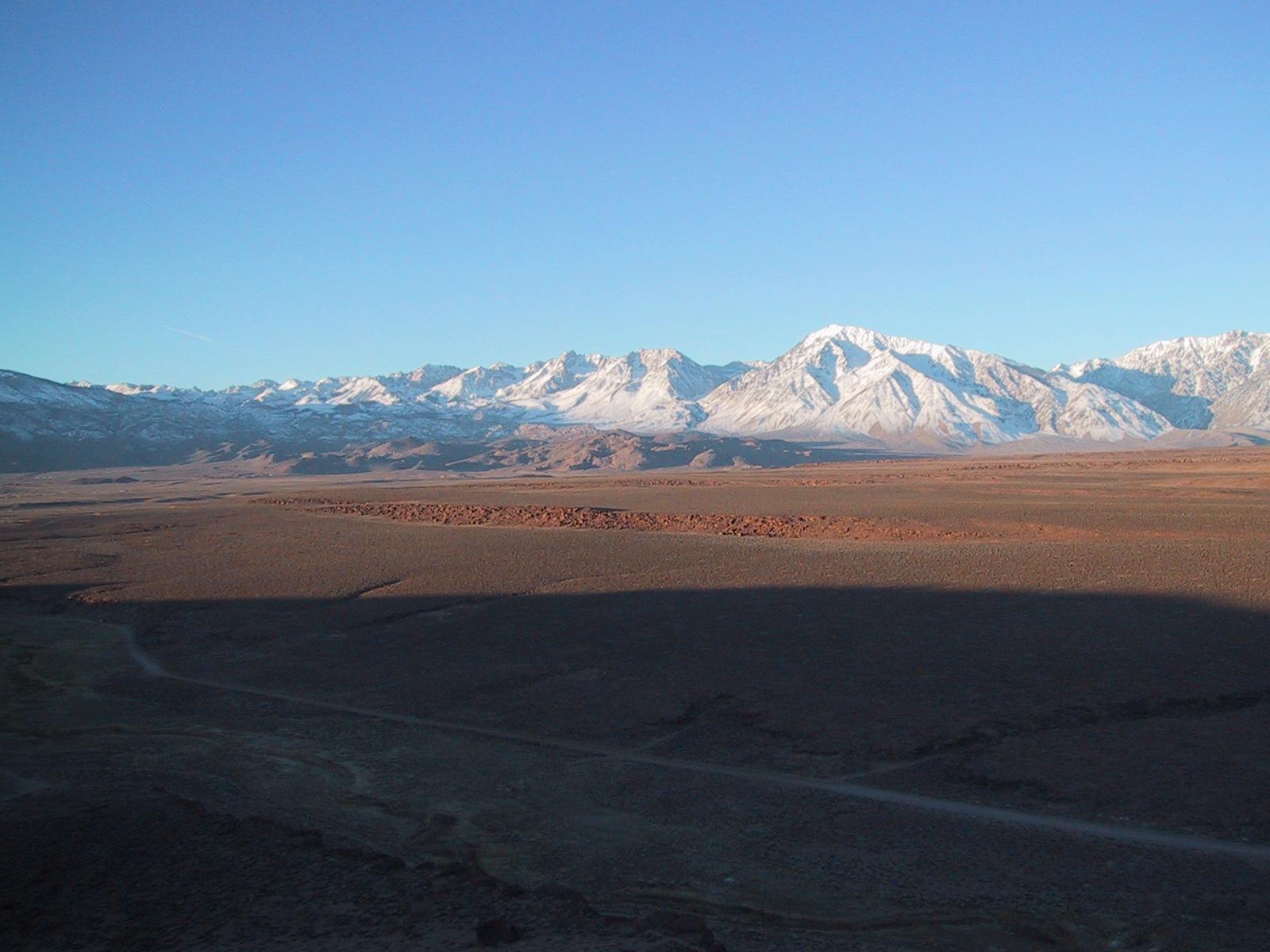Snow-capped mountains overlook a vast valley with long shadows under a clear sky.