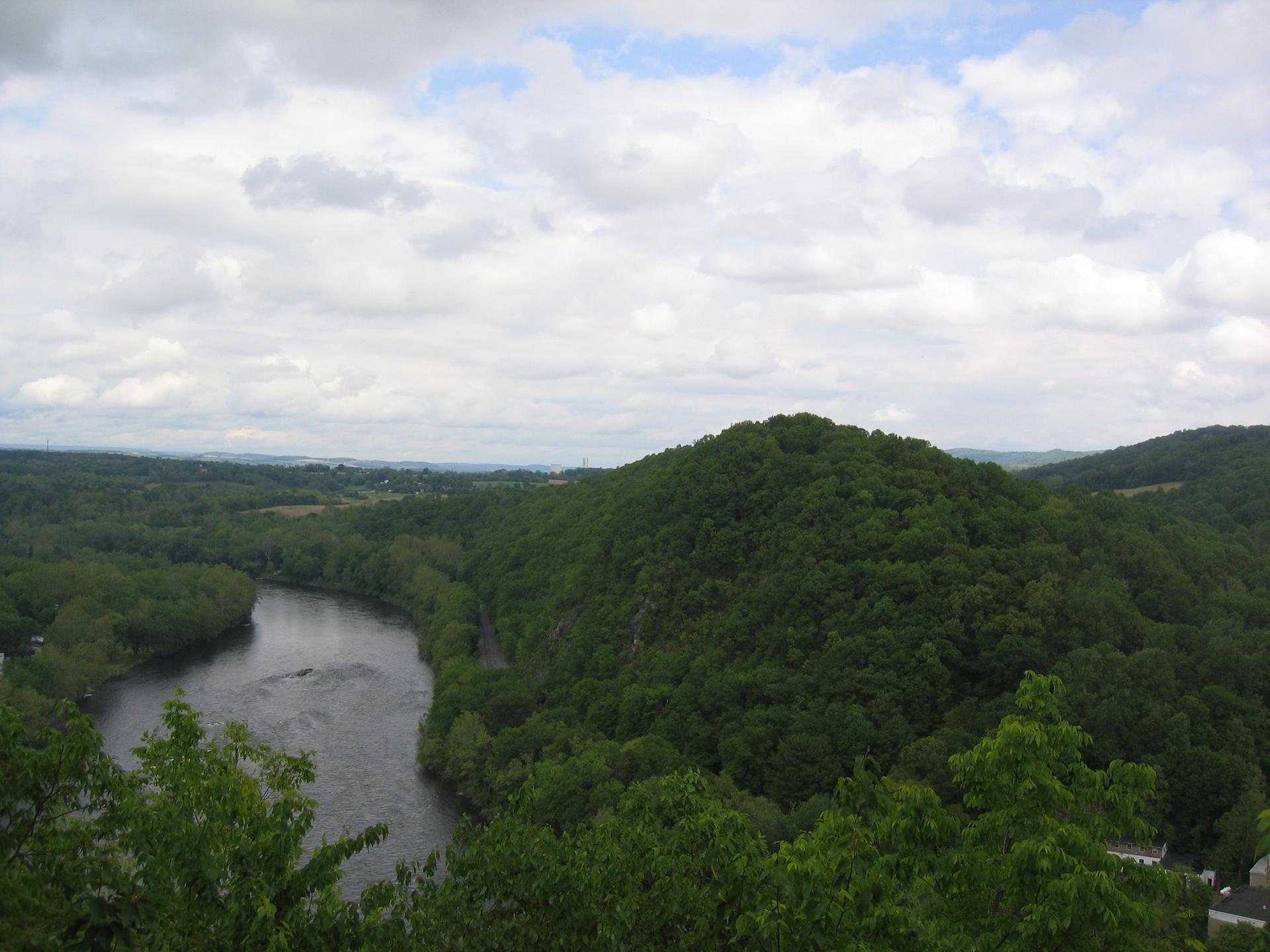 A wide river winds through lush green forested hills under a cloudy sky.