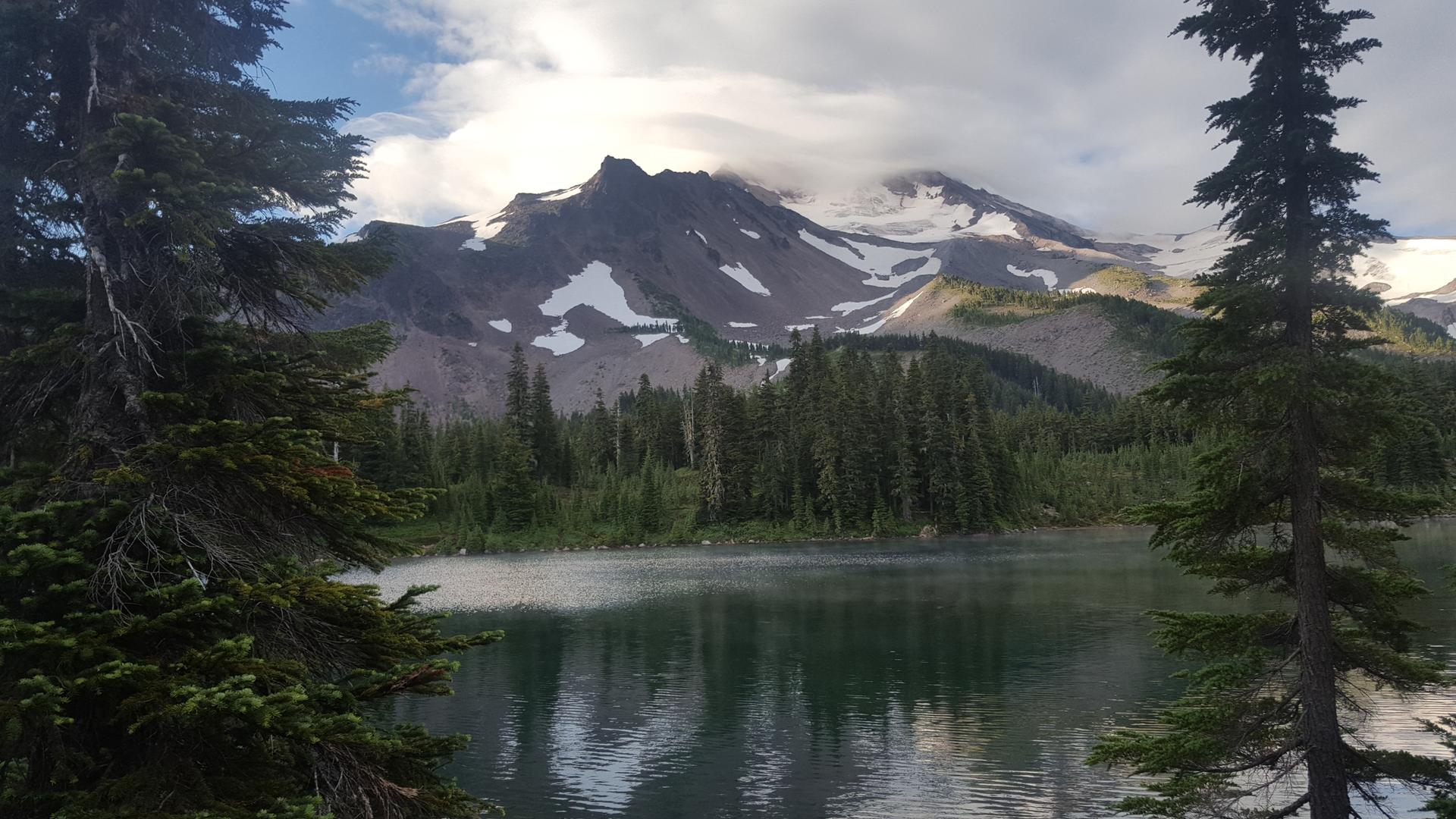 Serene lake reflecting a snow-capped mountain under cloudy skies, framed by trees.