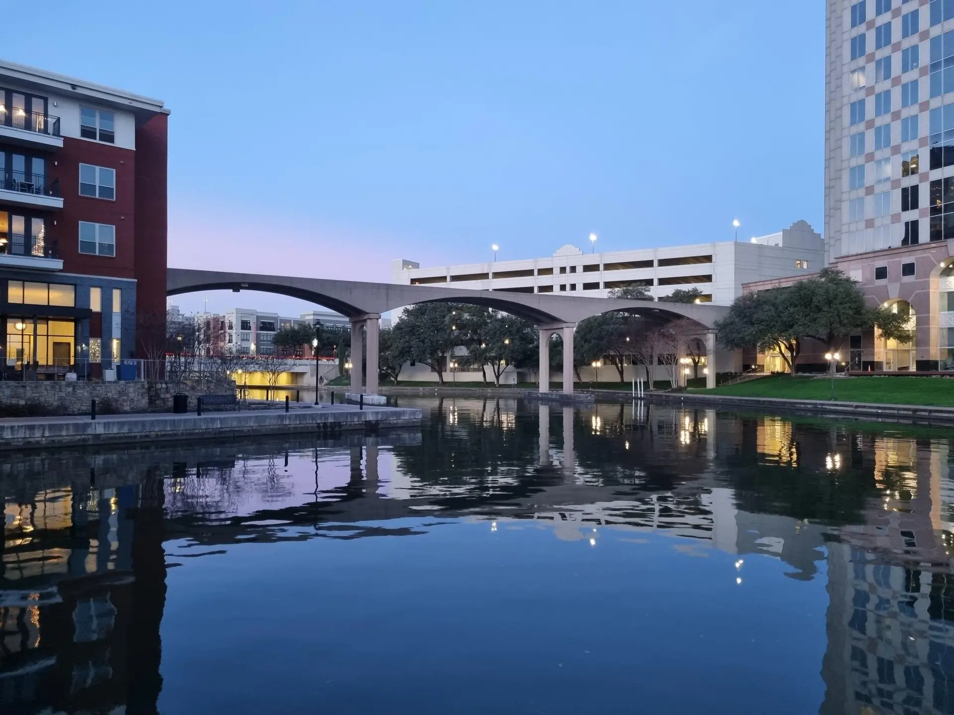 Evening view of a calm canal reflecting city buildings and an arched bridge.
