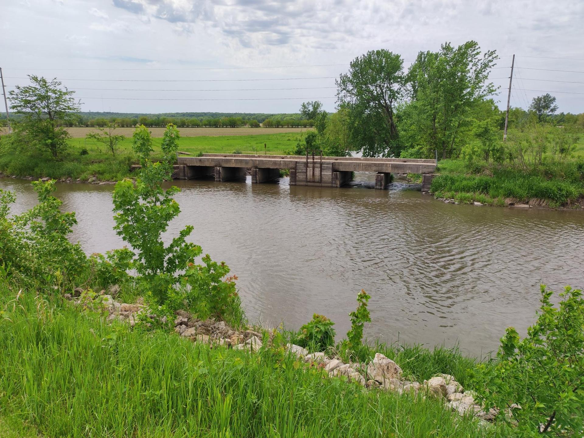 A concrete bridge spans a river with green banks under an overcast sky.