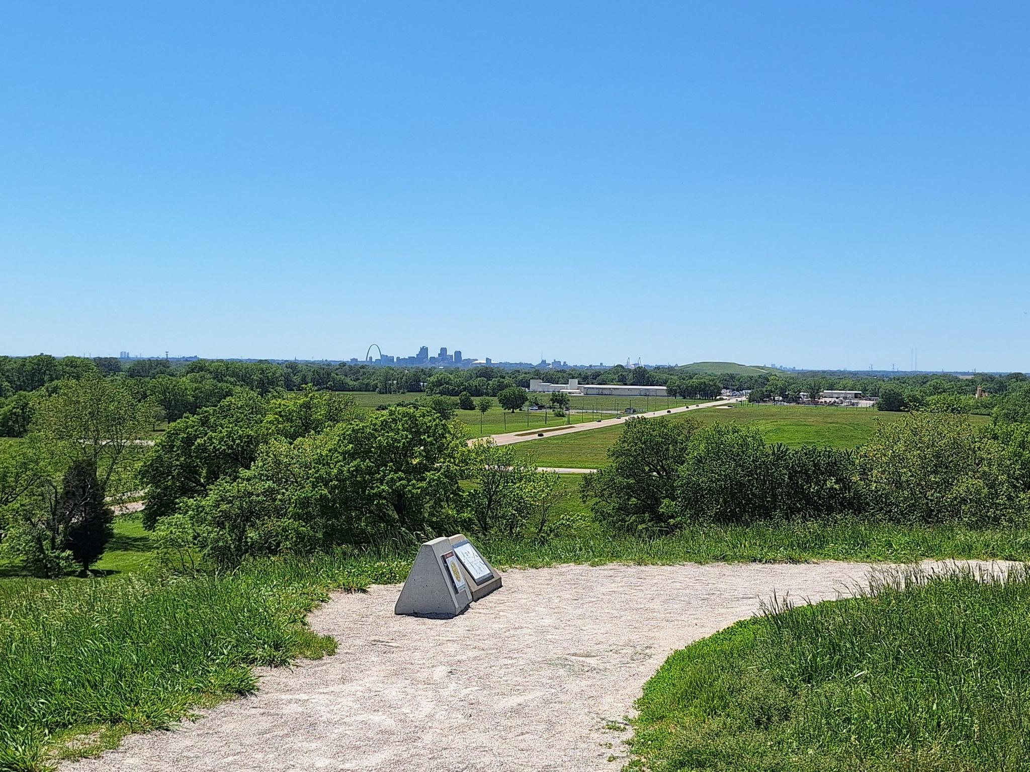 Sunny panoramic view from a hill with an informational sign and distant city.