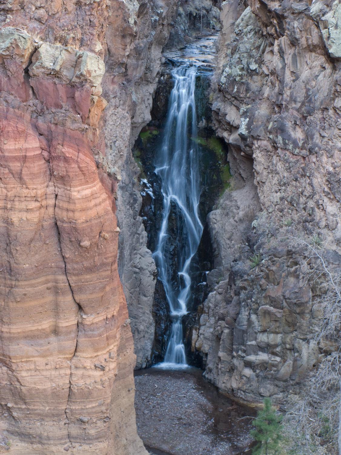 Tall waterfall cascades down a narrow rocky canyon with colorful rock formations.