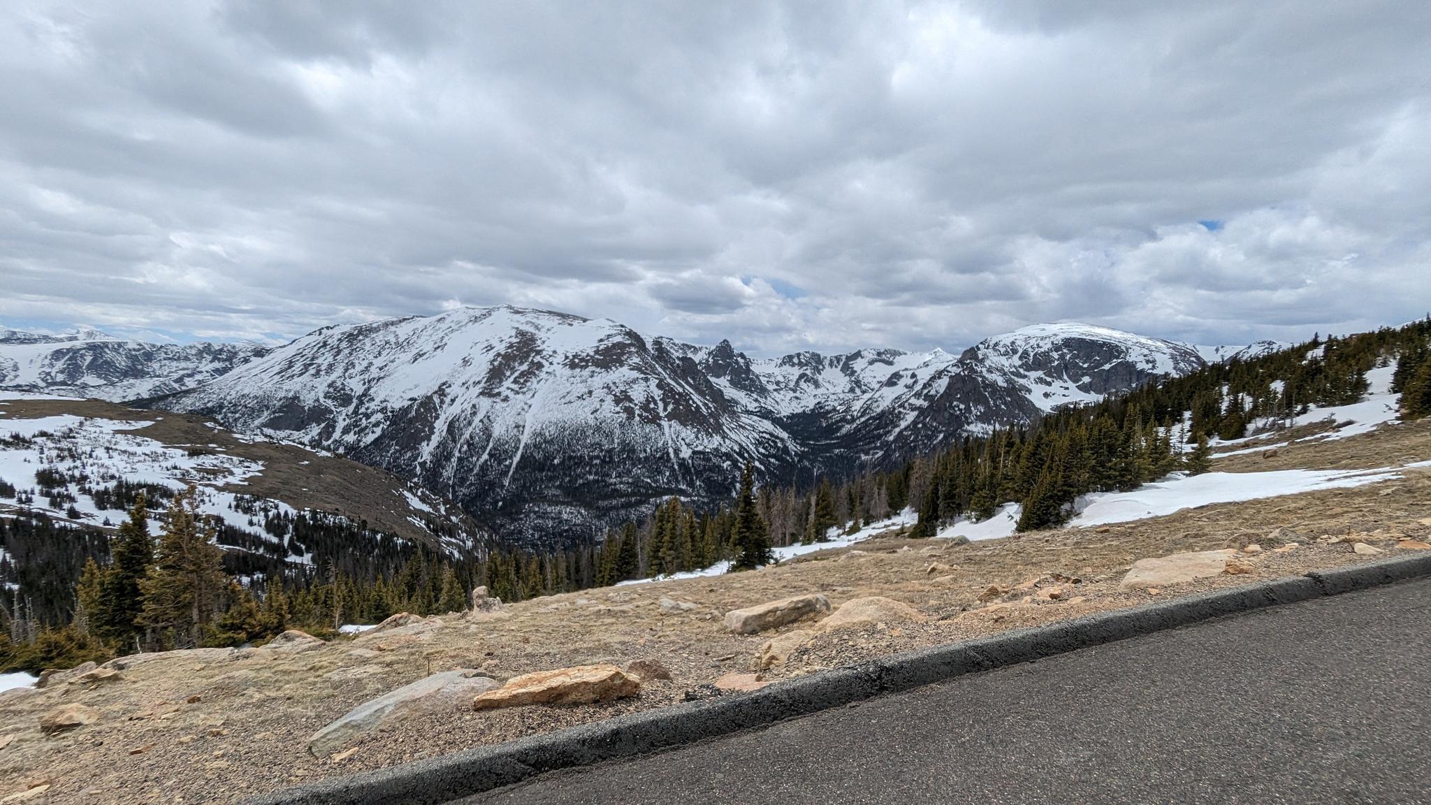 Snow-covered mountains and evergreen trees beside a road under an overcast sky.