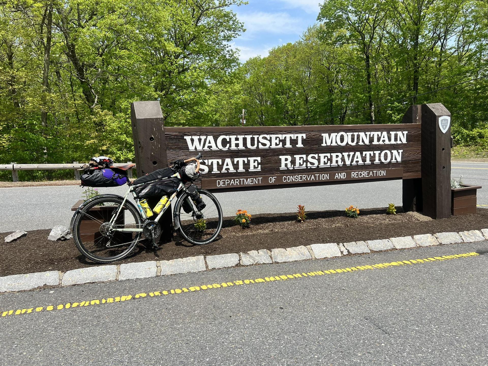 A touring bicycle with bags parked beside a large wooden sign on a sunny day.
