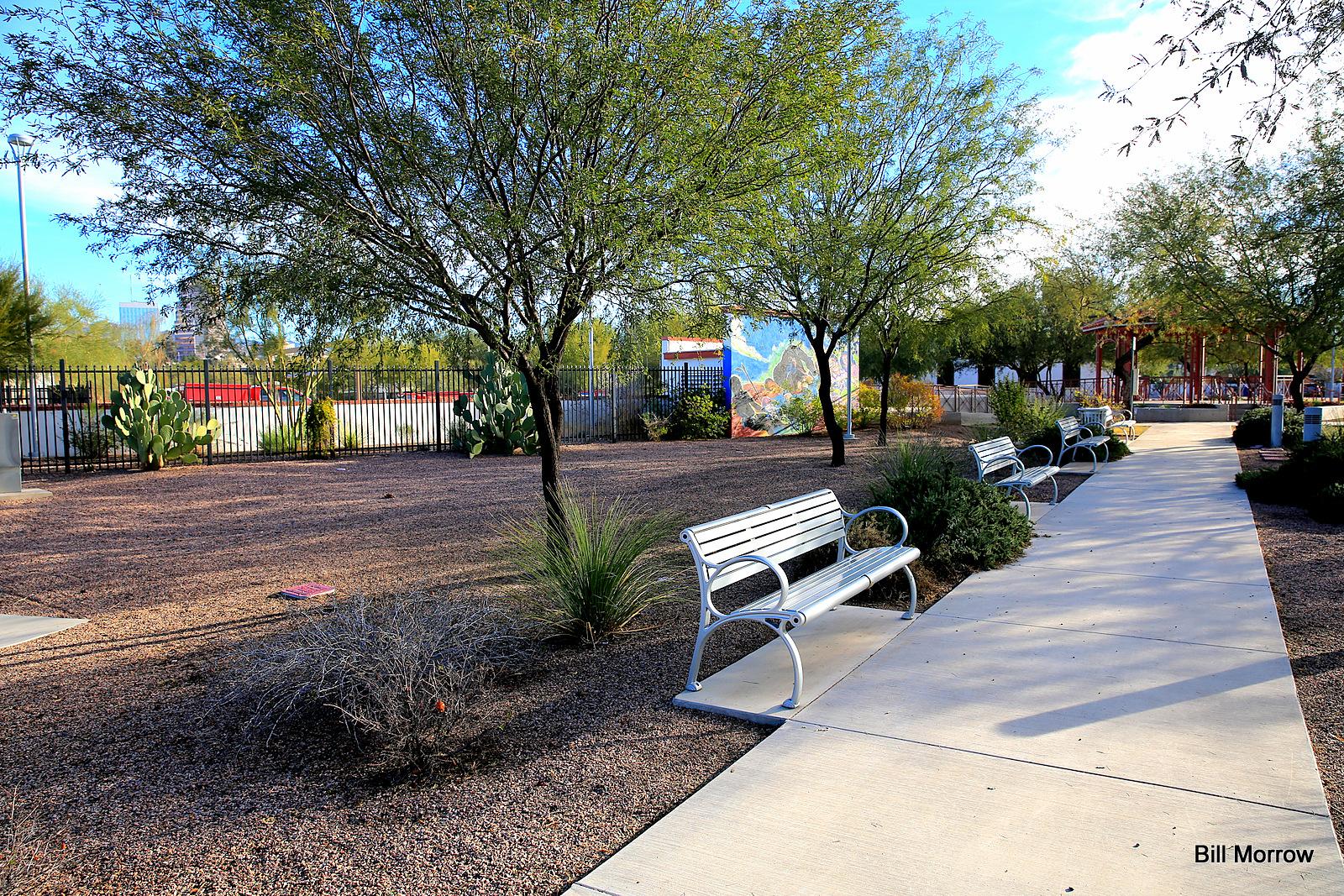 Sunny park with multiple benches along a paved path, surrounded by trees.