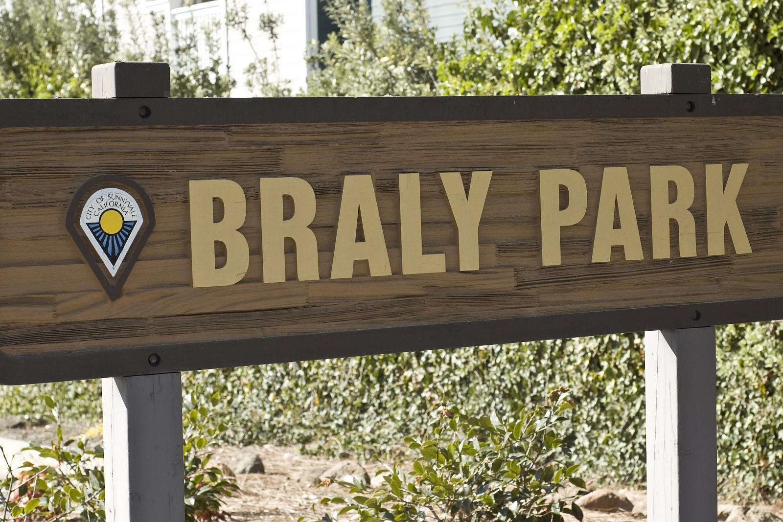 A wooden sign with gold letters stands against green foliage on a sunny day.