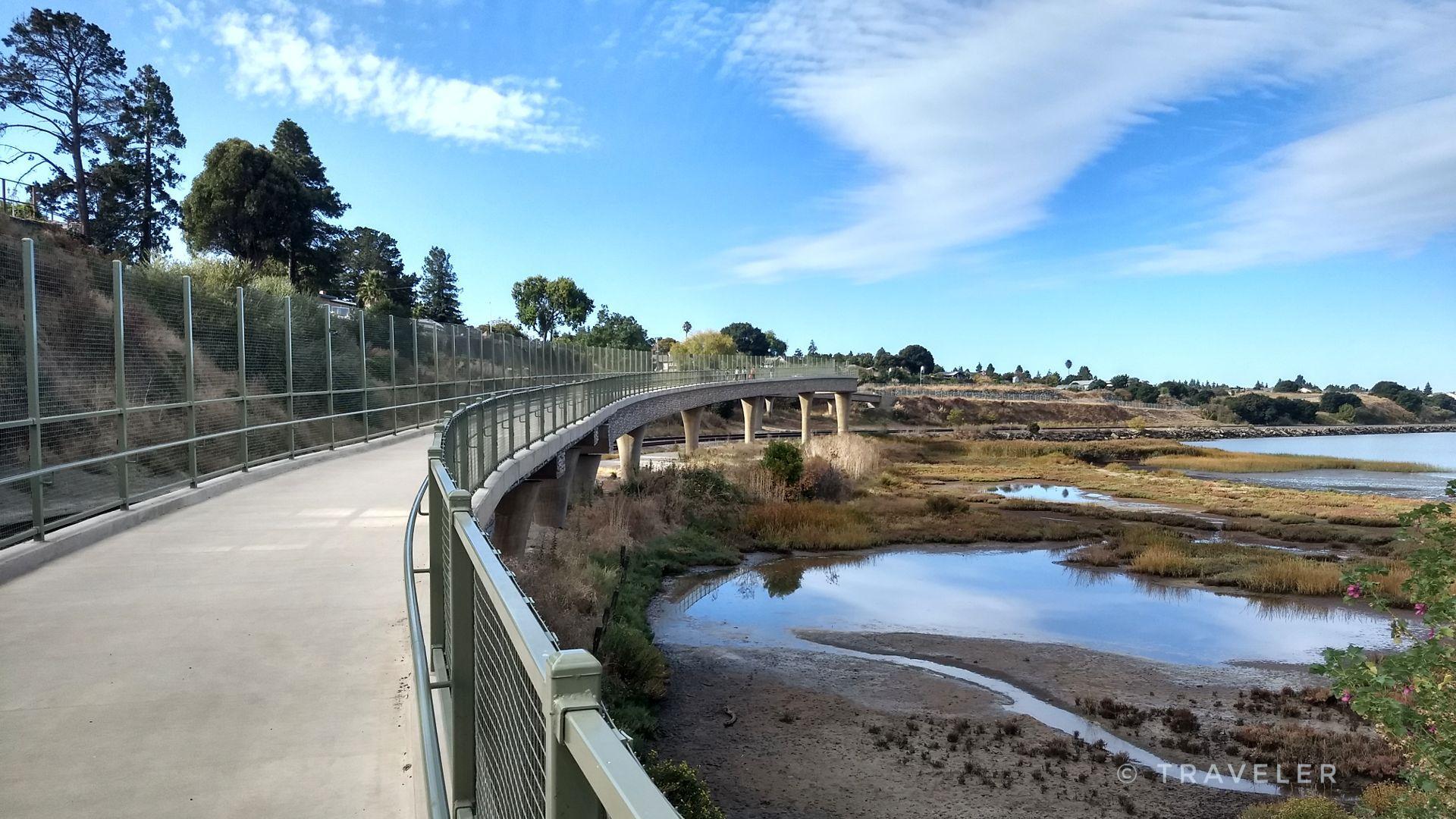 Curved concrete path with railing next to a marshy waterway under a blue sky.