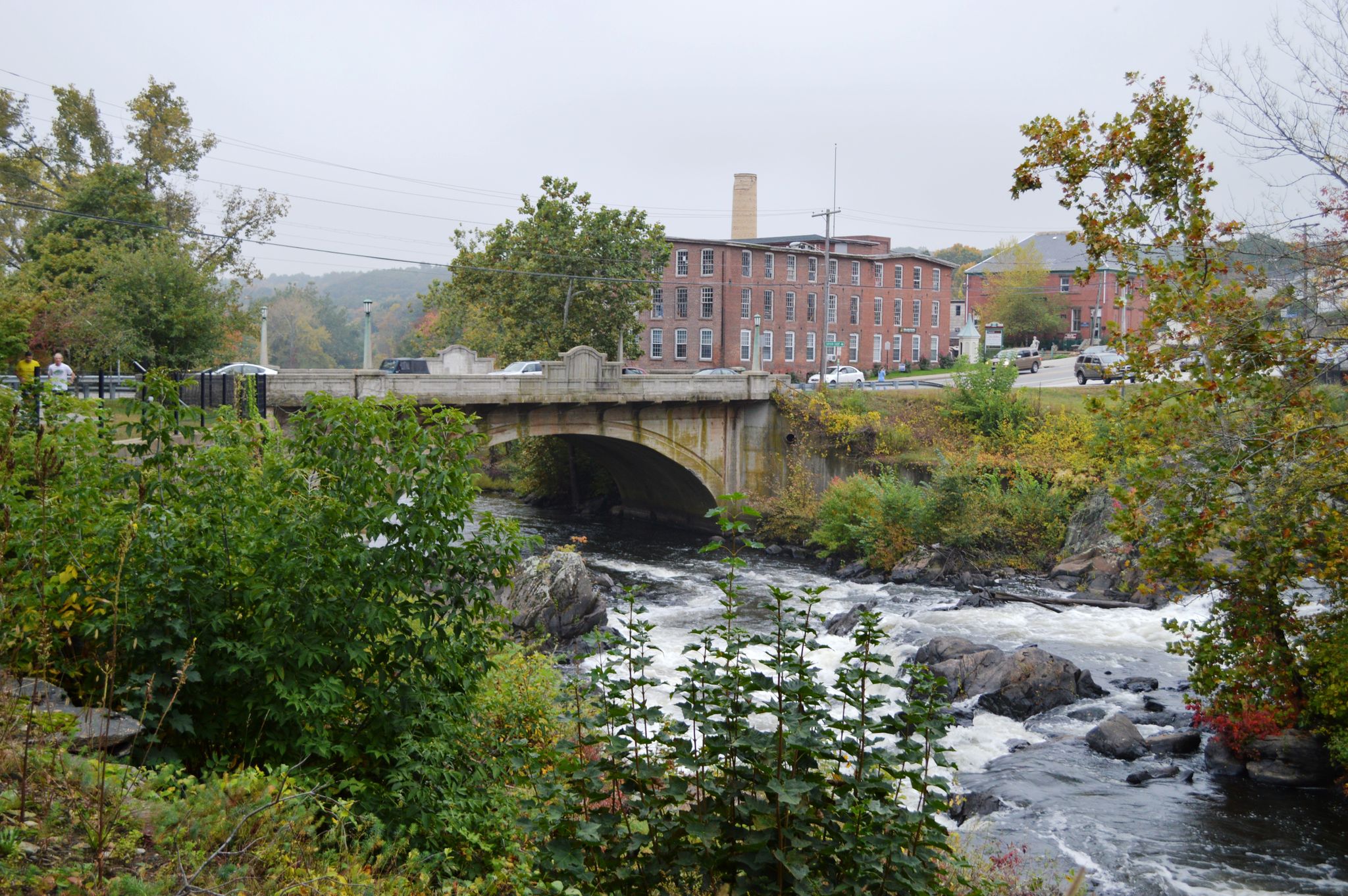 A stone arch bridge spans a rocky river with a large brick building. - Putnam River Trail