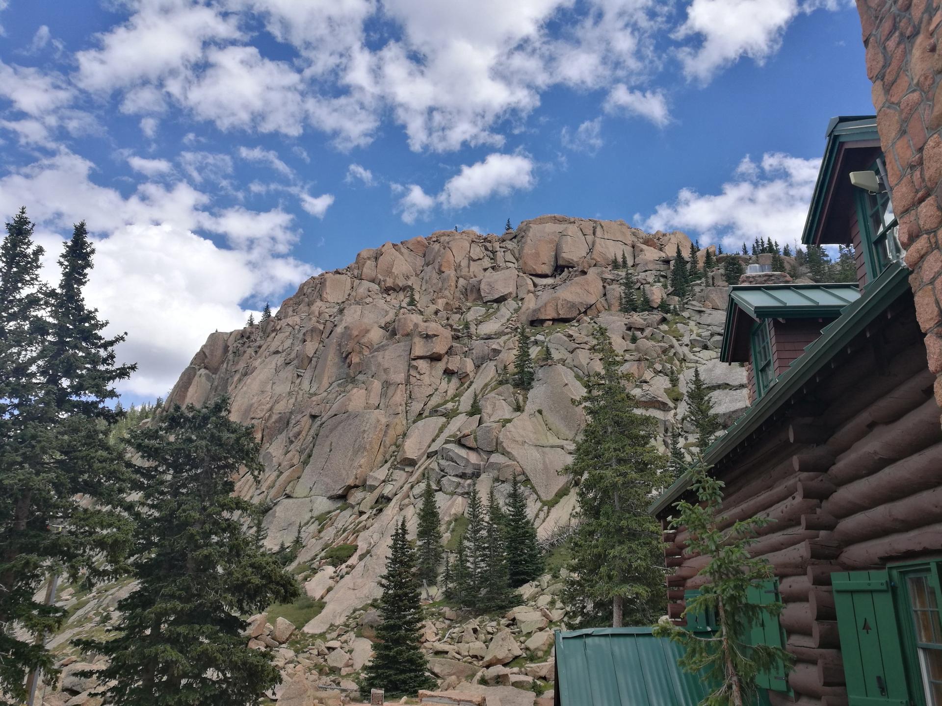 Massive rocky mountain face towering over a rustic log cabin and pine trees.