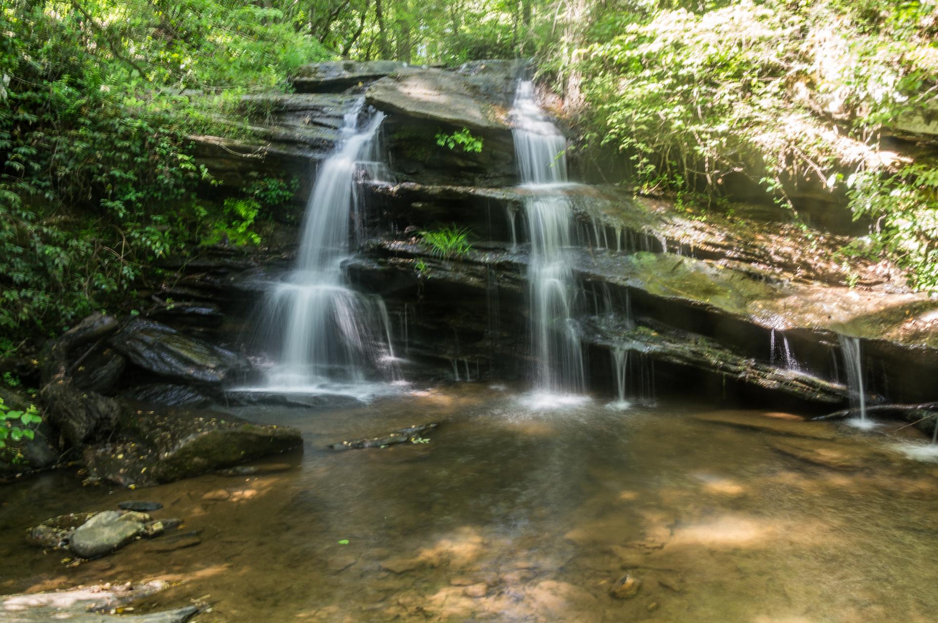 Multi-tiered waterfall cascading over dark rocks into a shallow pool in a sunny forest.