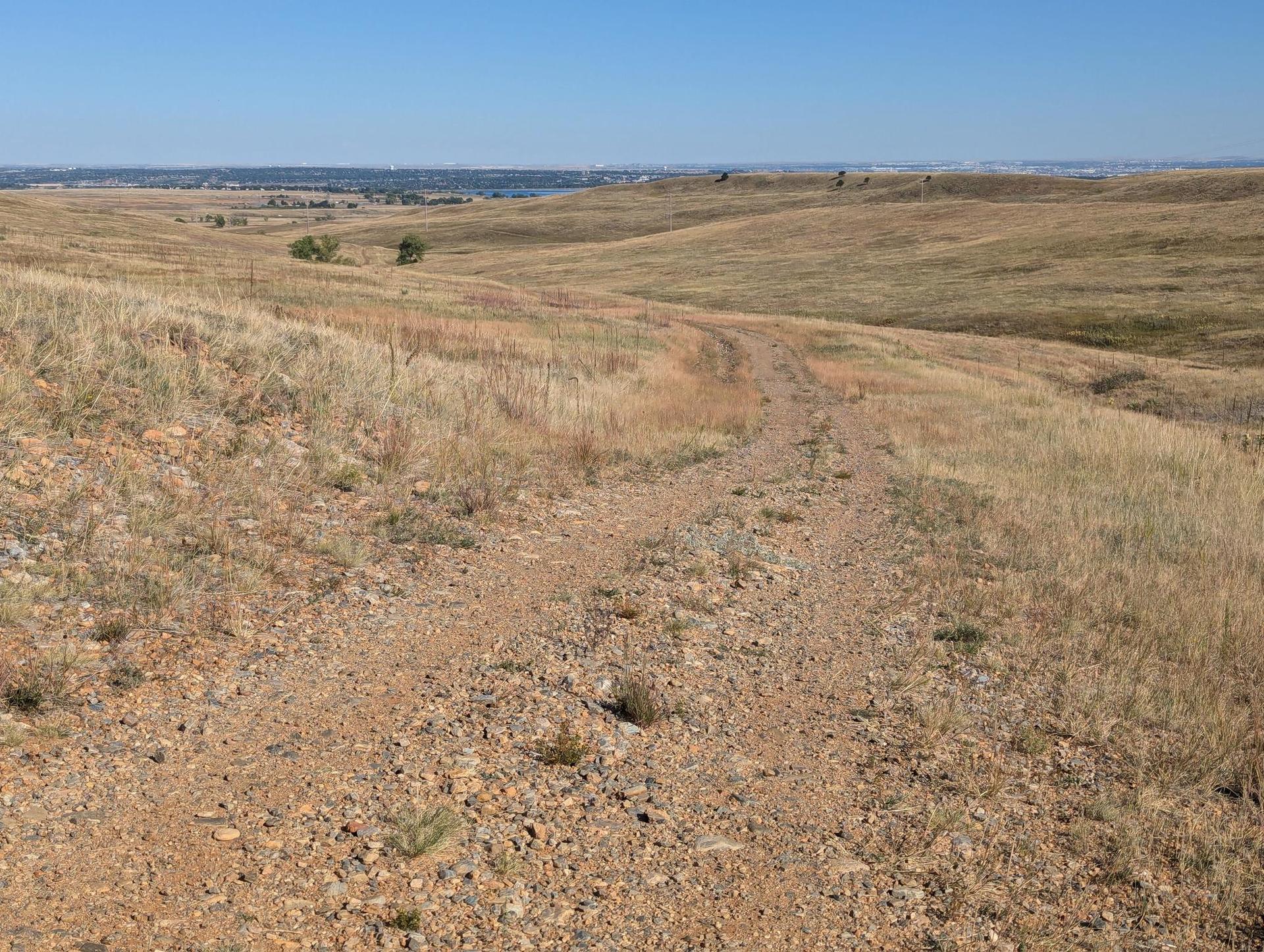 A dirt path winds through sunlit, dry, rolling hills under a clear sky.