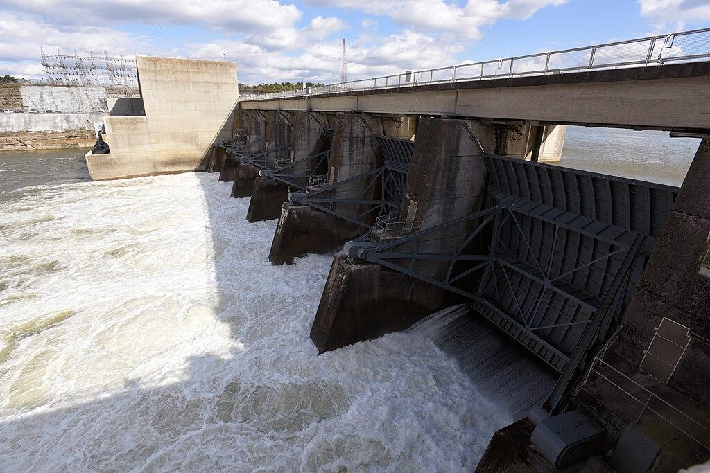 Large concrete dam with water rushing through multiple open gates under a cloudy sky.