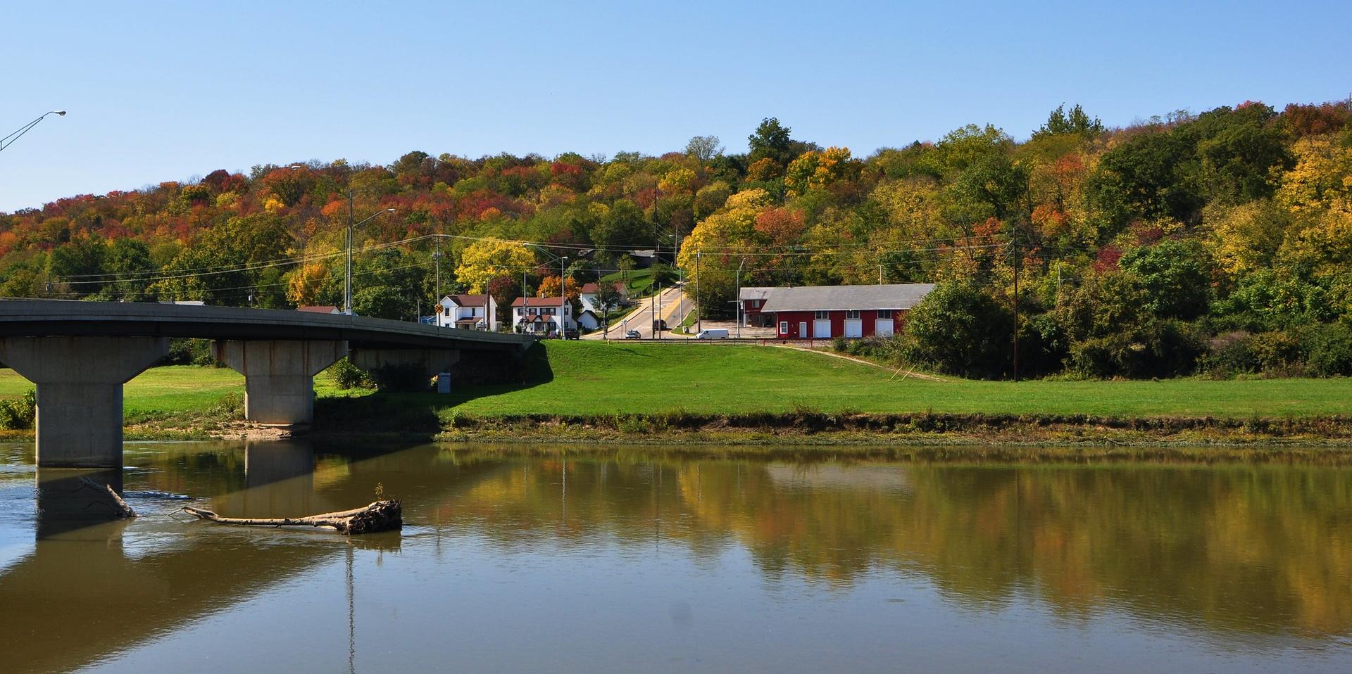 River reflecting autumn trees and a bridge, with buildings on a distant hill.