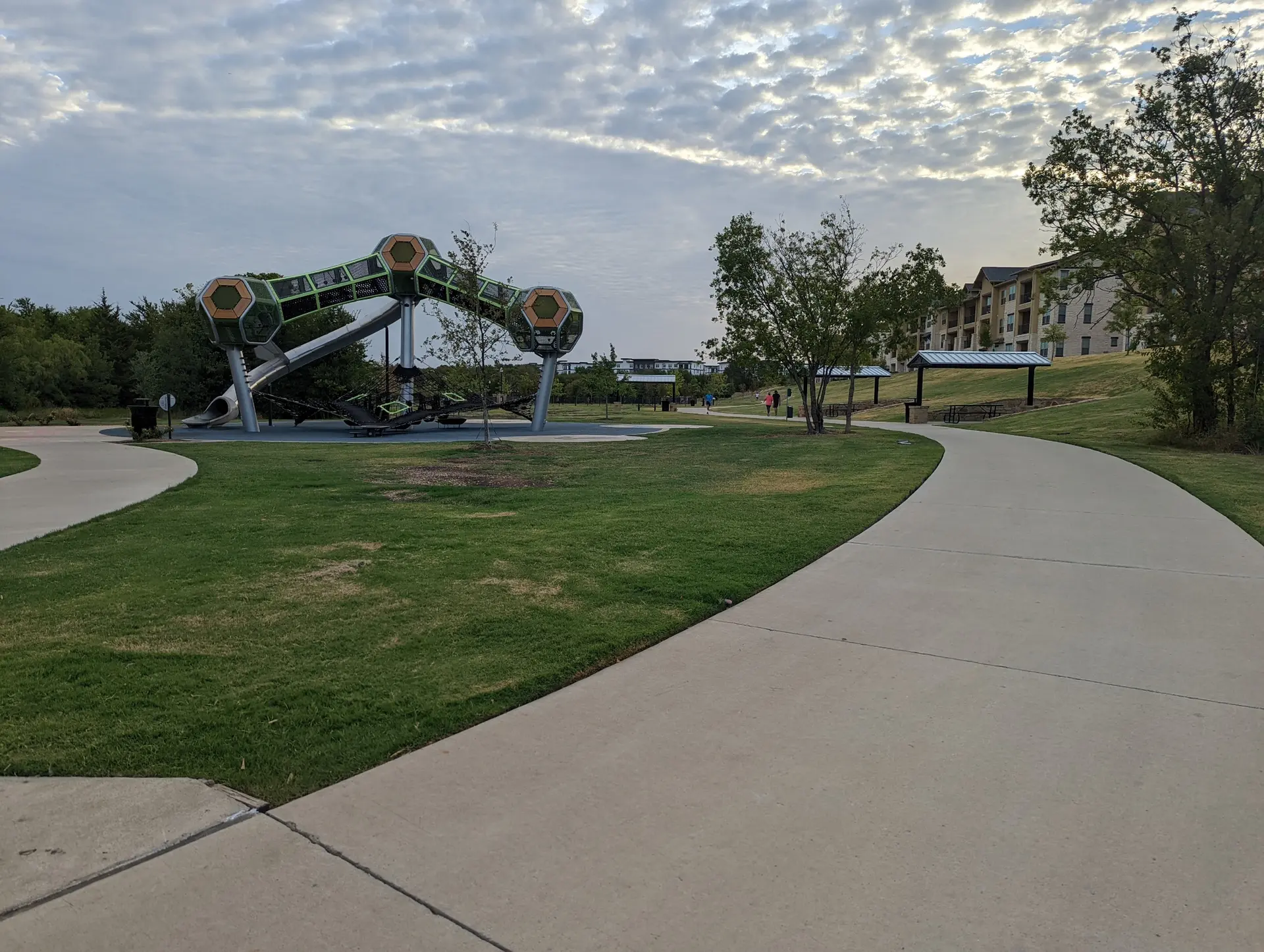 A paved path curves past a modern playground and grassy area under a cloudy sky.