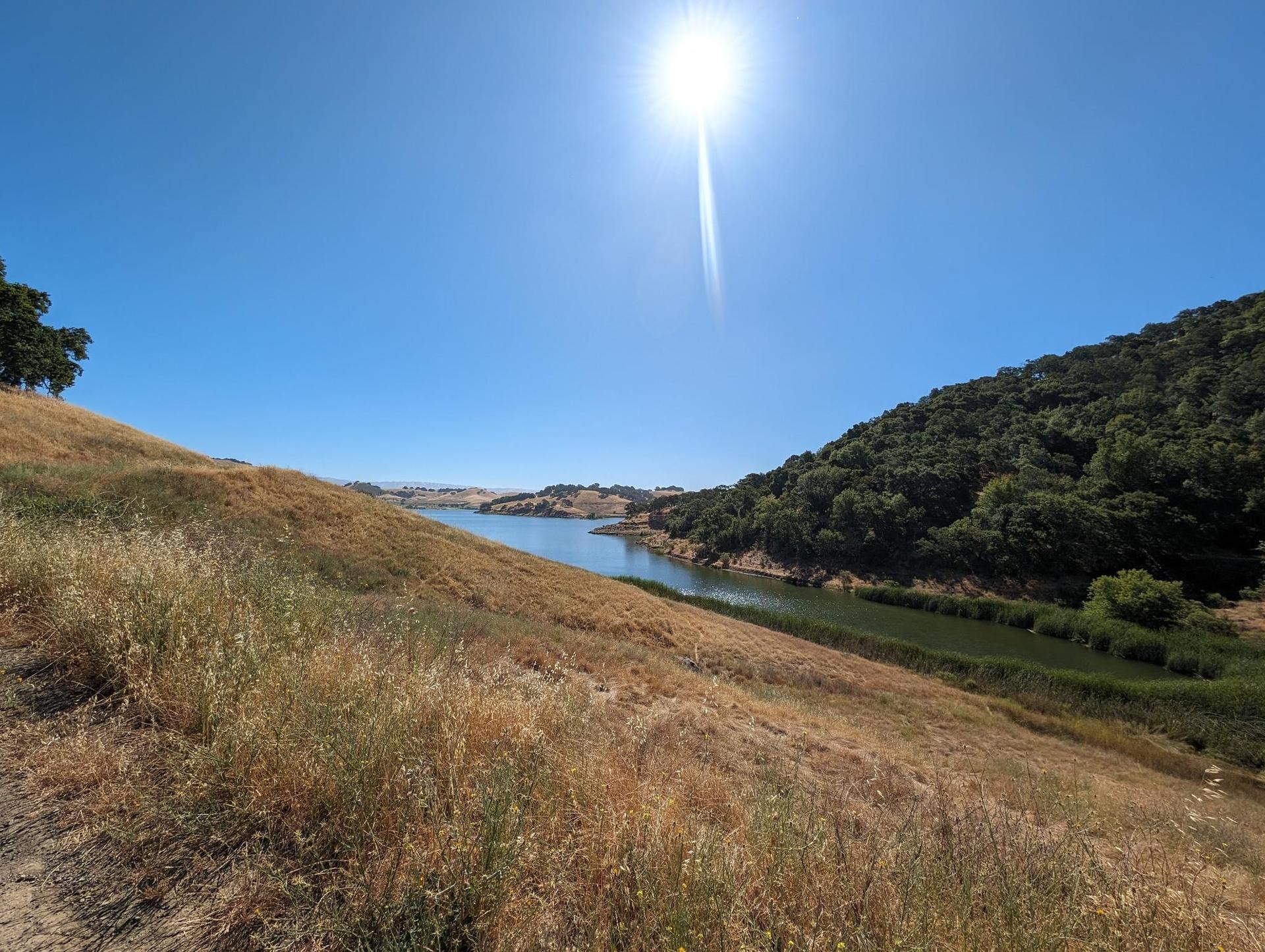 Bright sun over a lake nestled between dry grassy and green wooded hills.