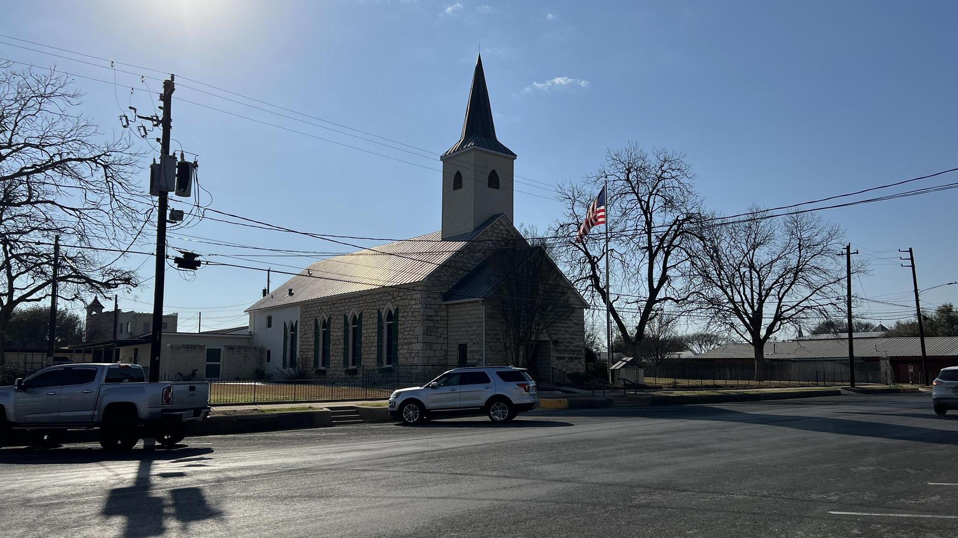 A stone church with a steeple, bare trees, and cars on a sunny street.
