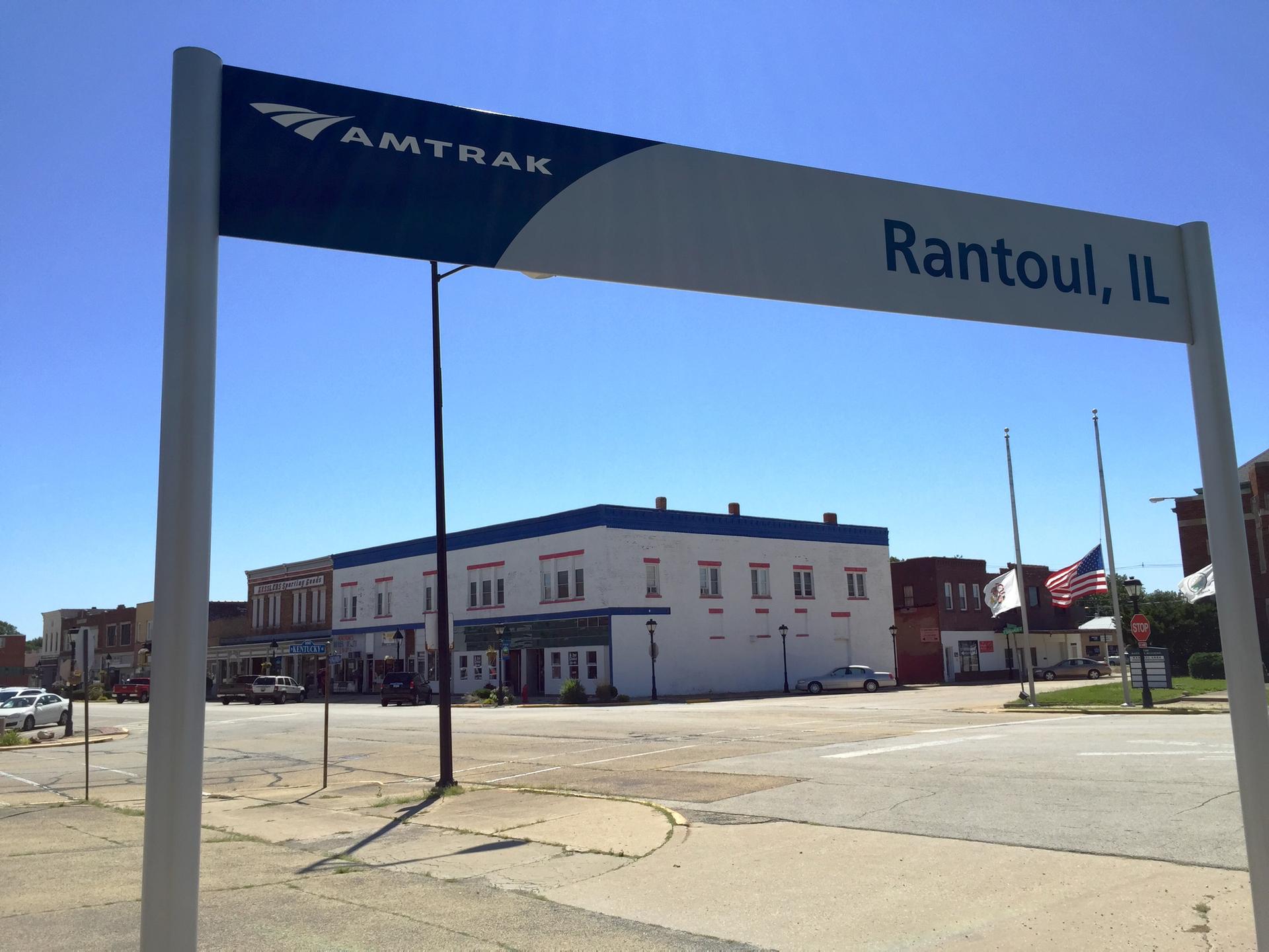 A large sign with a train logo overlooks a sunny street intersection and buildings.