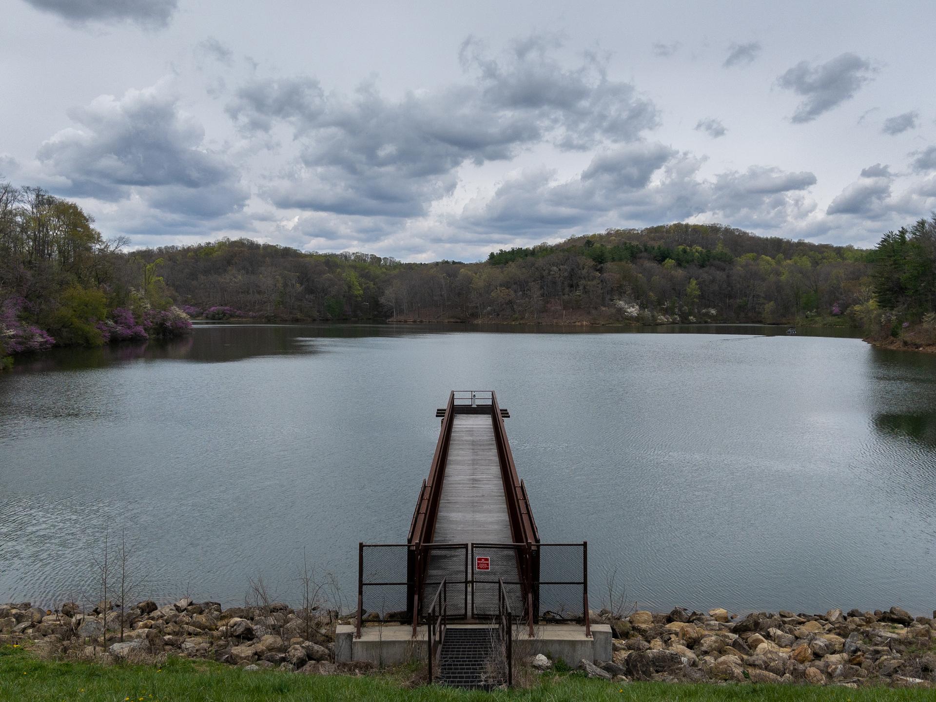 A long pier extends into a calm lake surrounded by forested hills under a cloudy sky.