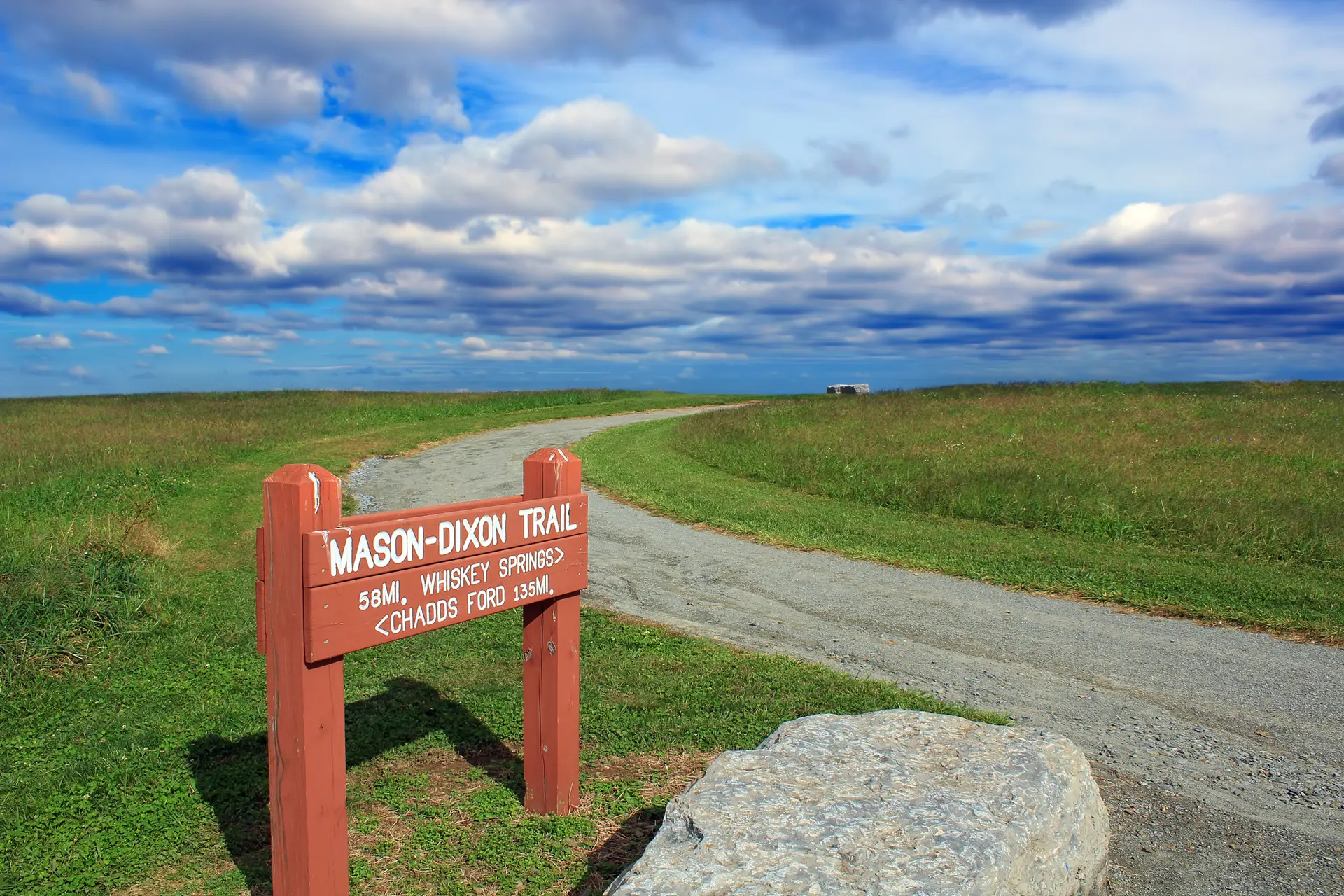A red wooden sign marks a winding dirt path through a sunny, open field.