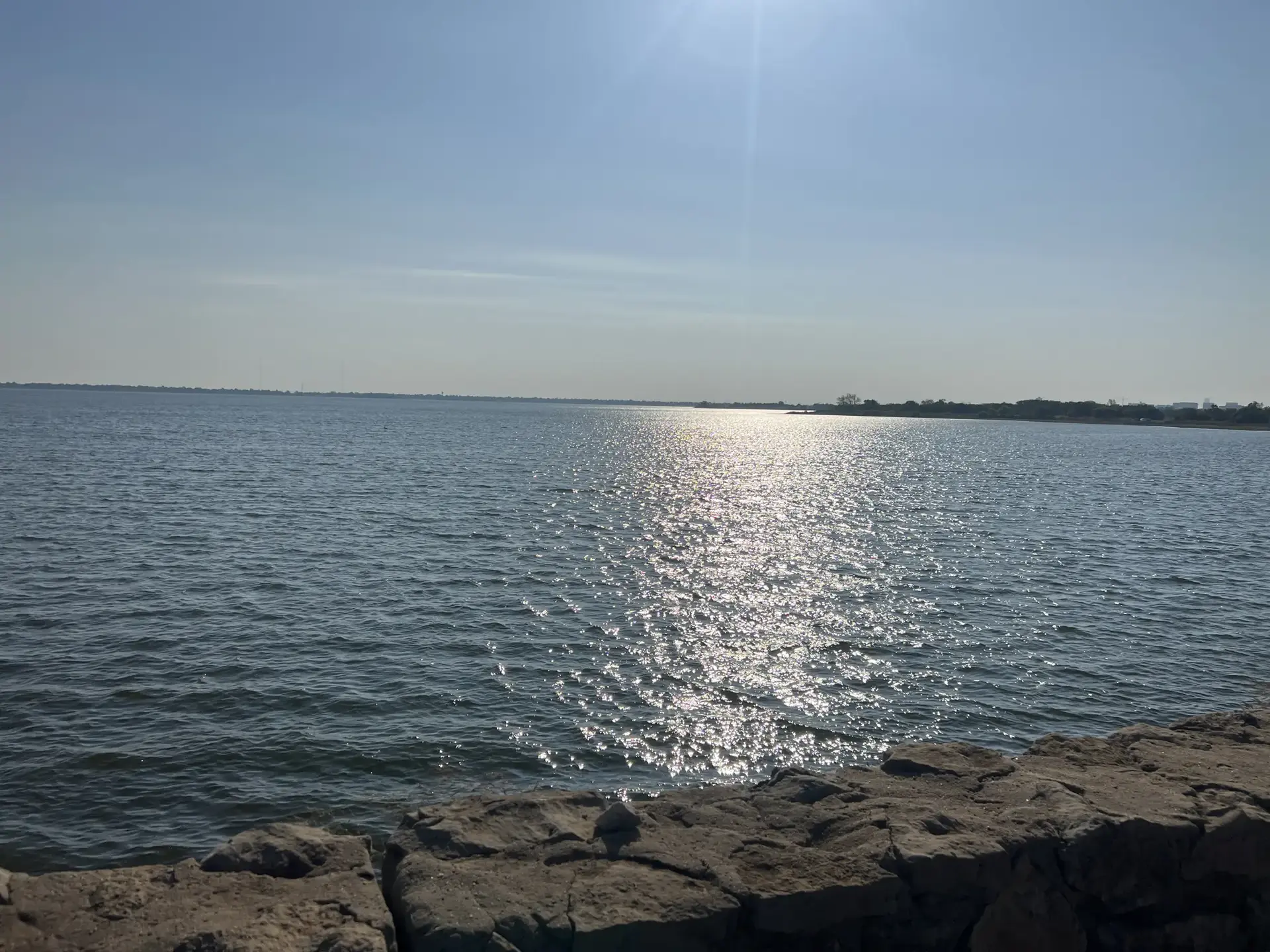 Bright sun reflects on a vast body of water with rocky foreground.
