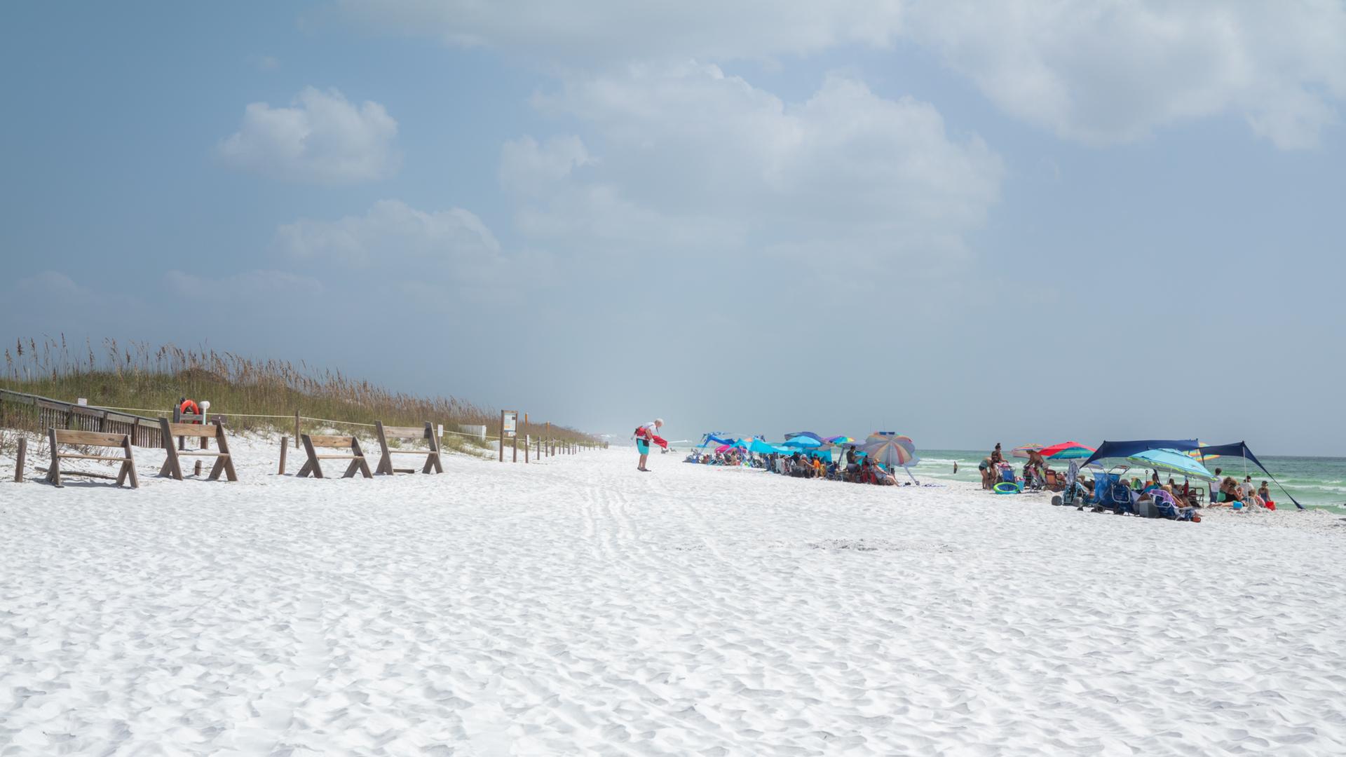 Crowded white sand beach with colorful umbrellas and people under an overcast sky.