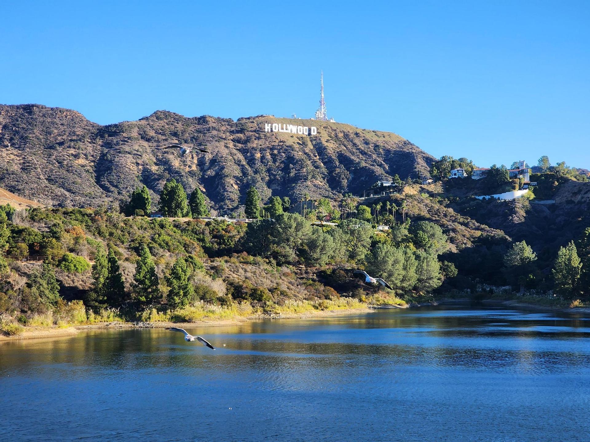 A clear blue lake with green trees and dry hills under bright skies.