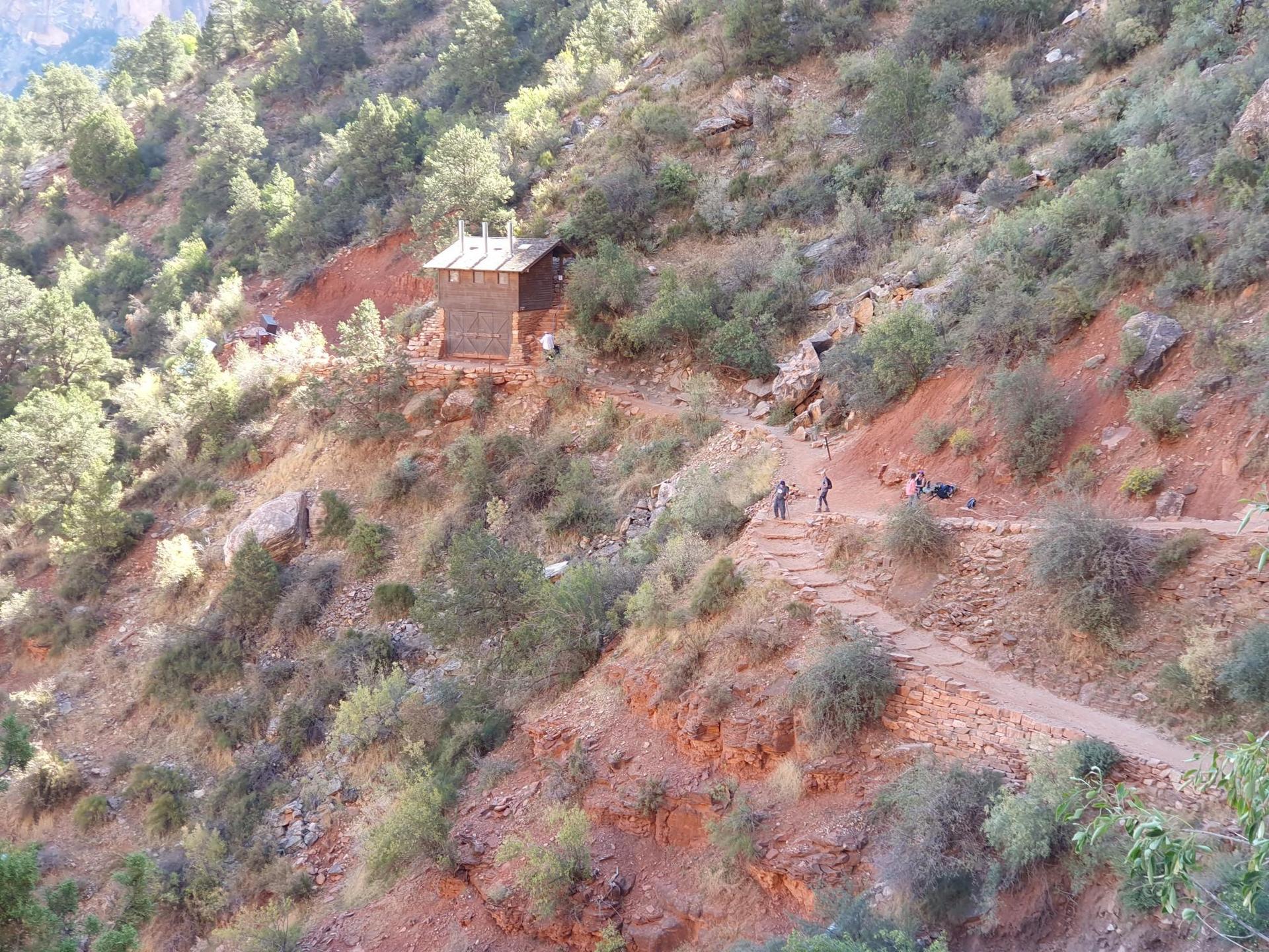 People on a winding dirt trail on a sunny hillside with a small building.