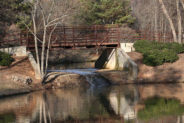 A metal pedestrian bridge crosses a small stream with trees and bushes.