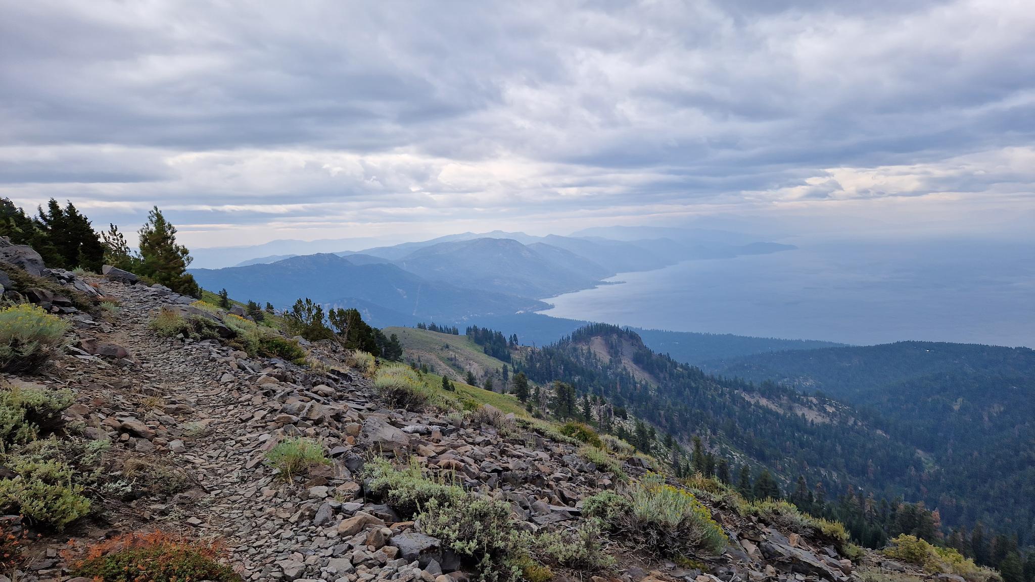 Rocky mountain trail overlooking a vast lake and distant peaks under cloudy skies.