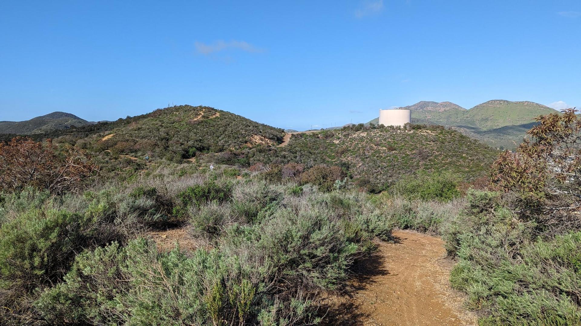 A dirt path winds through sunny, scrub-covered hills with a water tank in the distance.