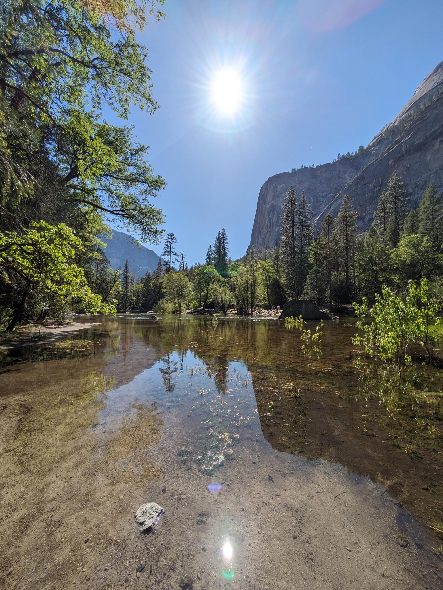 Sun shines brightly over a calm lake reflecting green trees and towering cliffs.