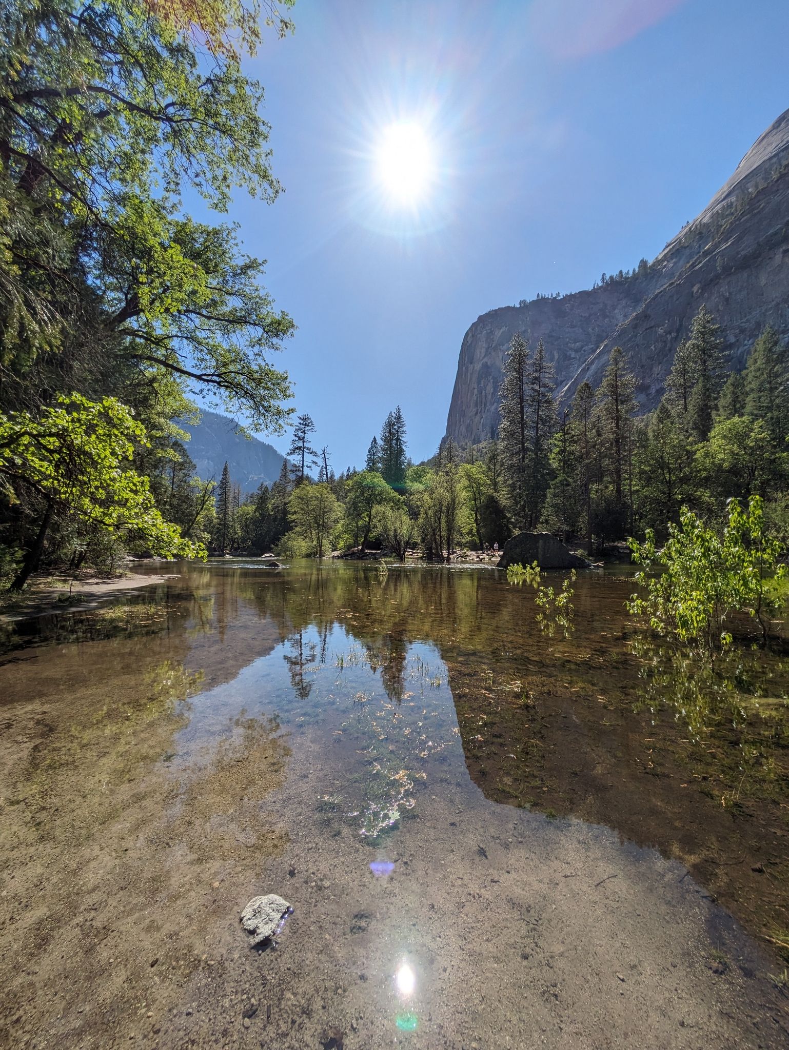 Sun shines brightly over a calm lake reflecting green trees and towering cliffs. - Yosemite Valley Loop Trail