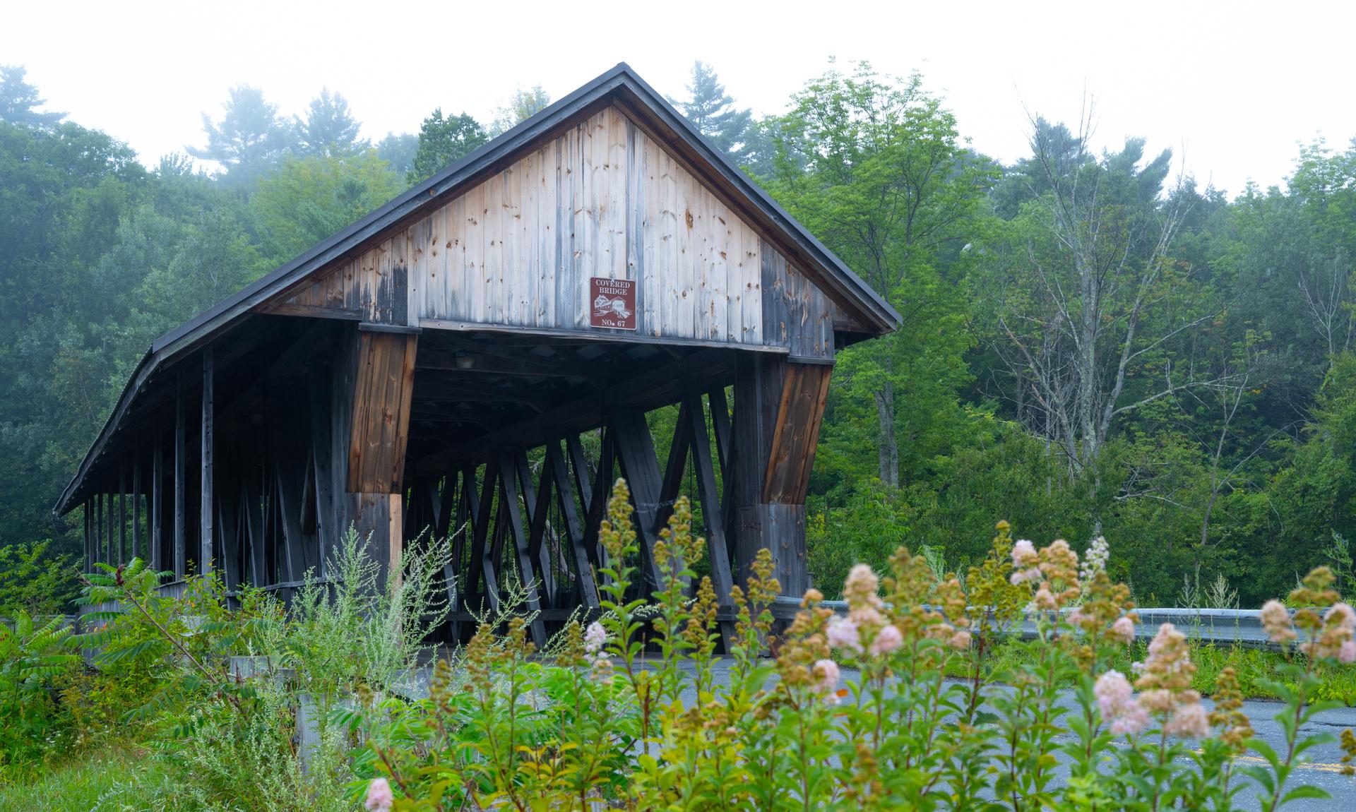 Rustic wooden covered bridge surrounded by green trees and wildflowers.