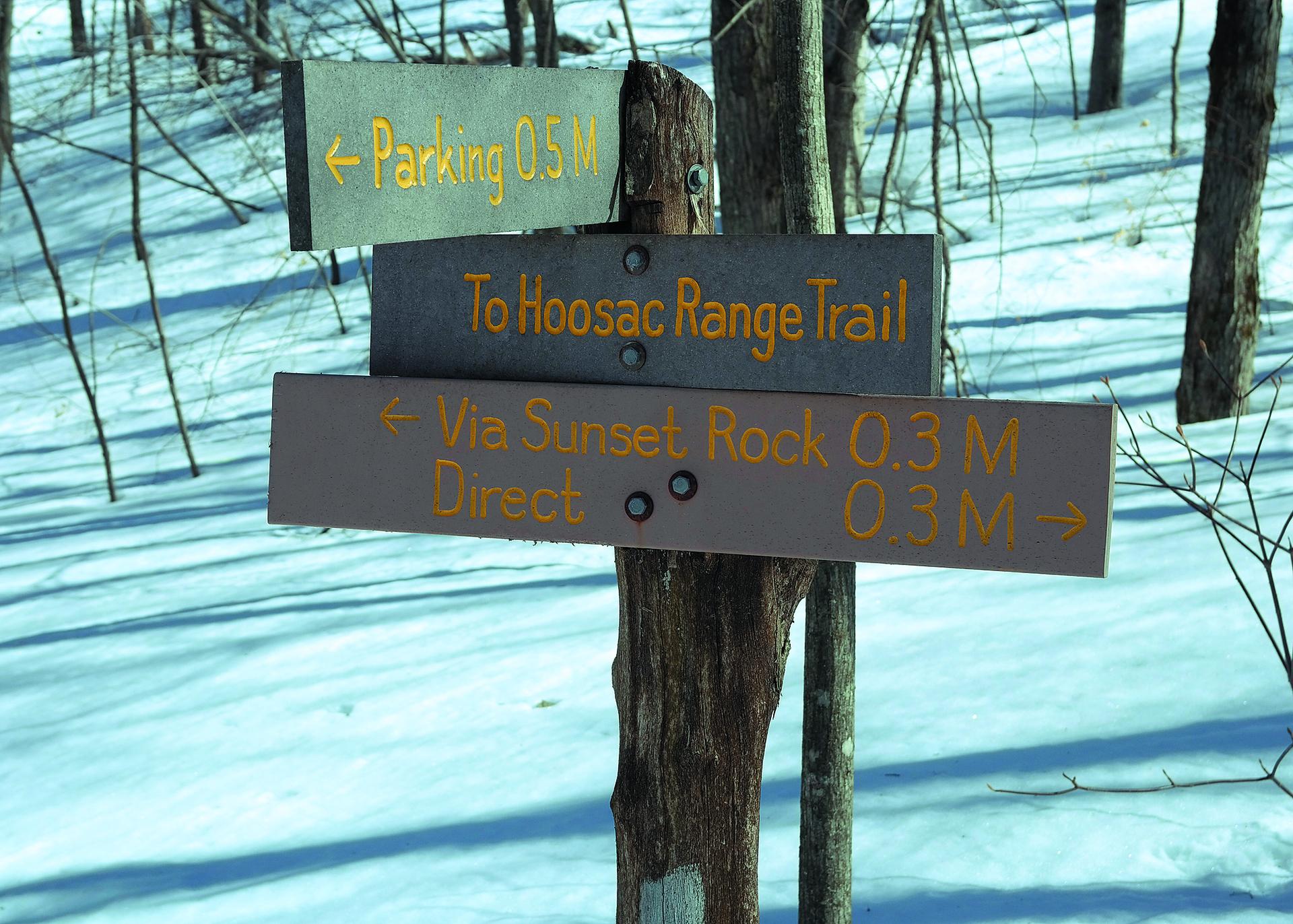 Wooden trail signs in a snowy forest, pointing to parking and trails.