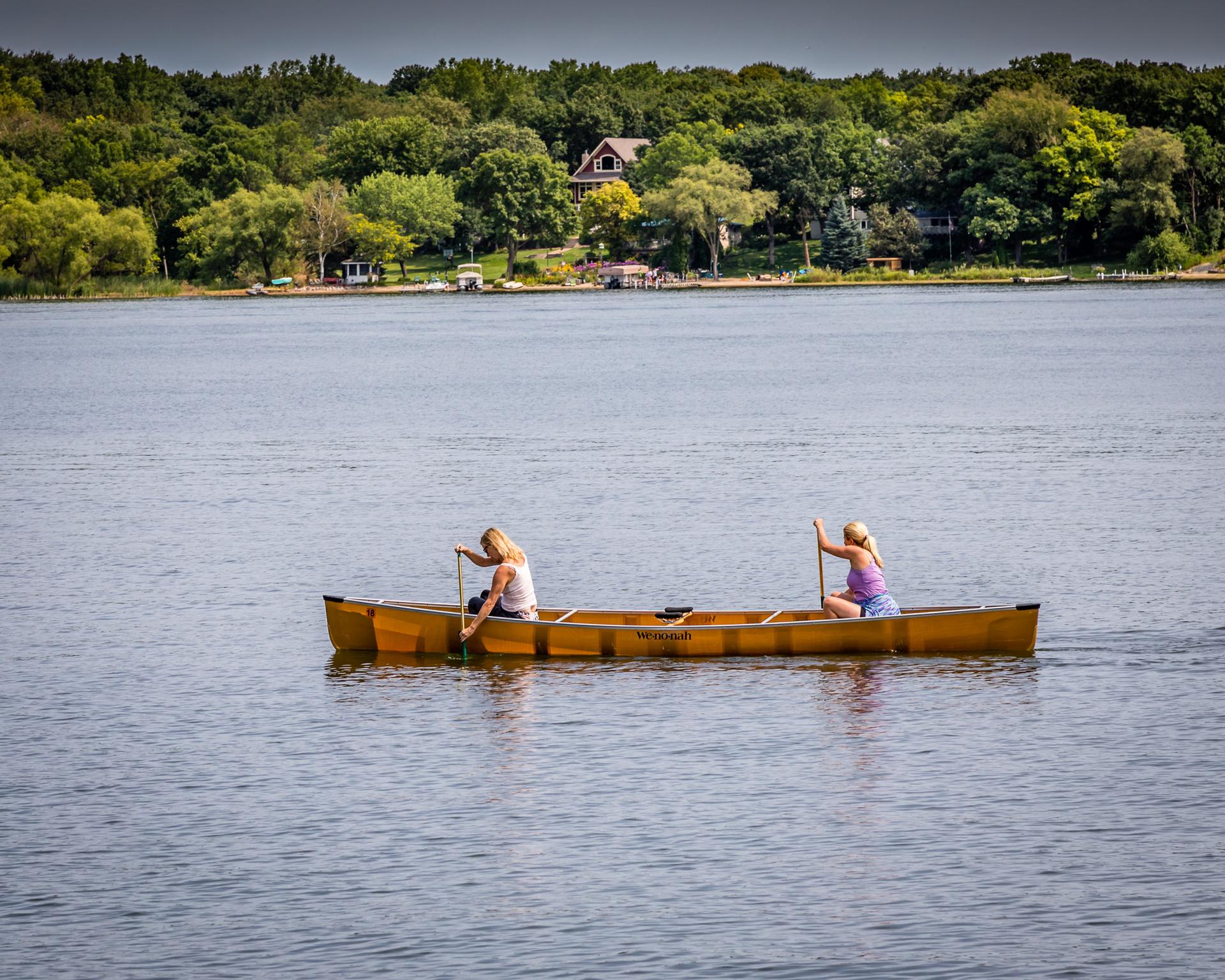 Two people paddle a yellow canoe on a calm lake under an overcast sky.