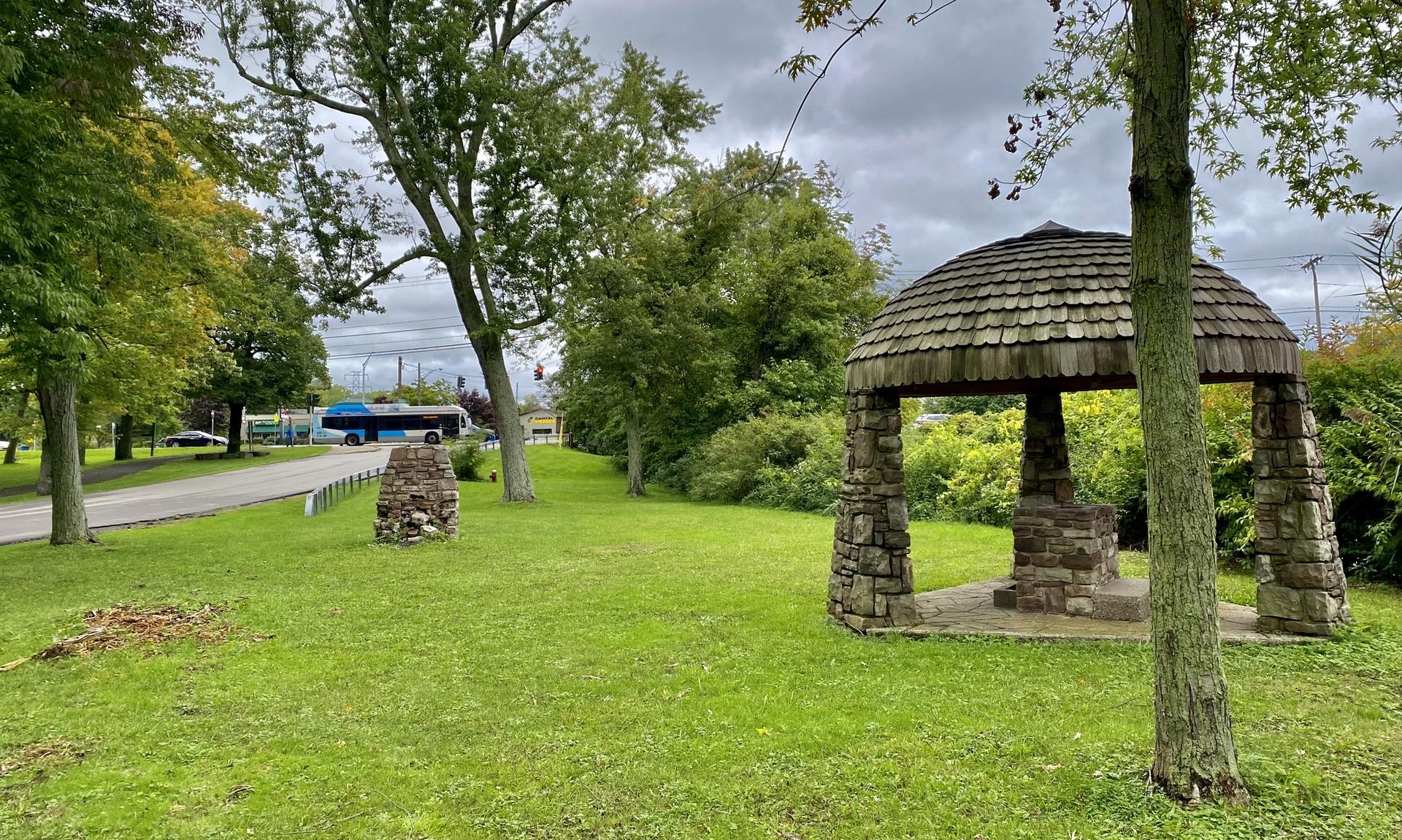 A stone gazebo and grassy park under an overcast sky, with a bus in the background.
