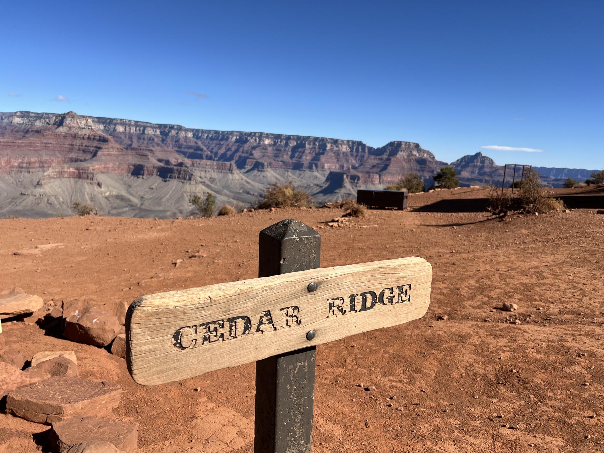 Wooden sign in foreground with a vast, sunlit canyon landscape behind.