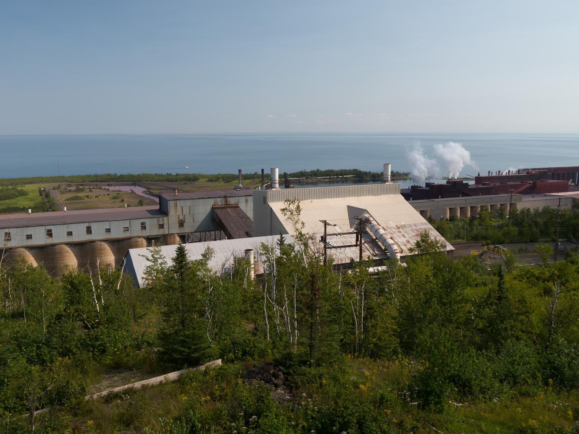 Industrial buildings with smoke plumes overlook a vast blue body of water.