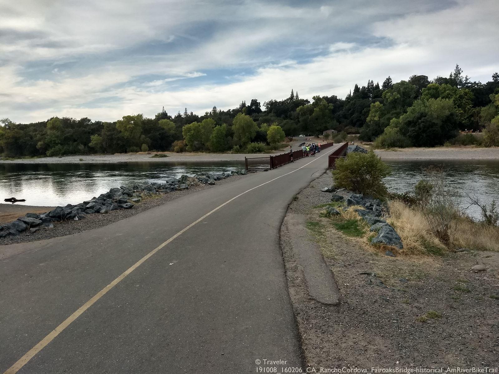 Paved path alongside a river leading to a bridge with cyclists under cloudy skies.