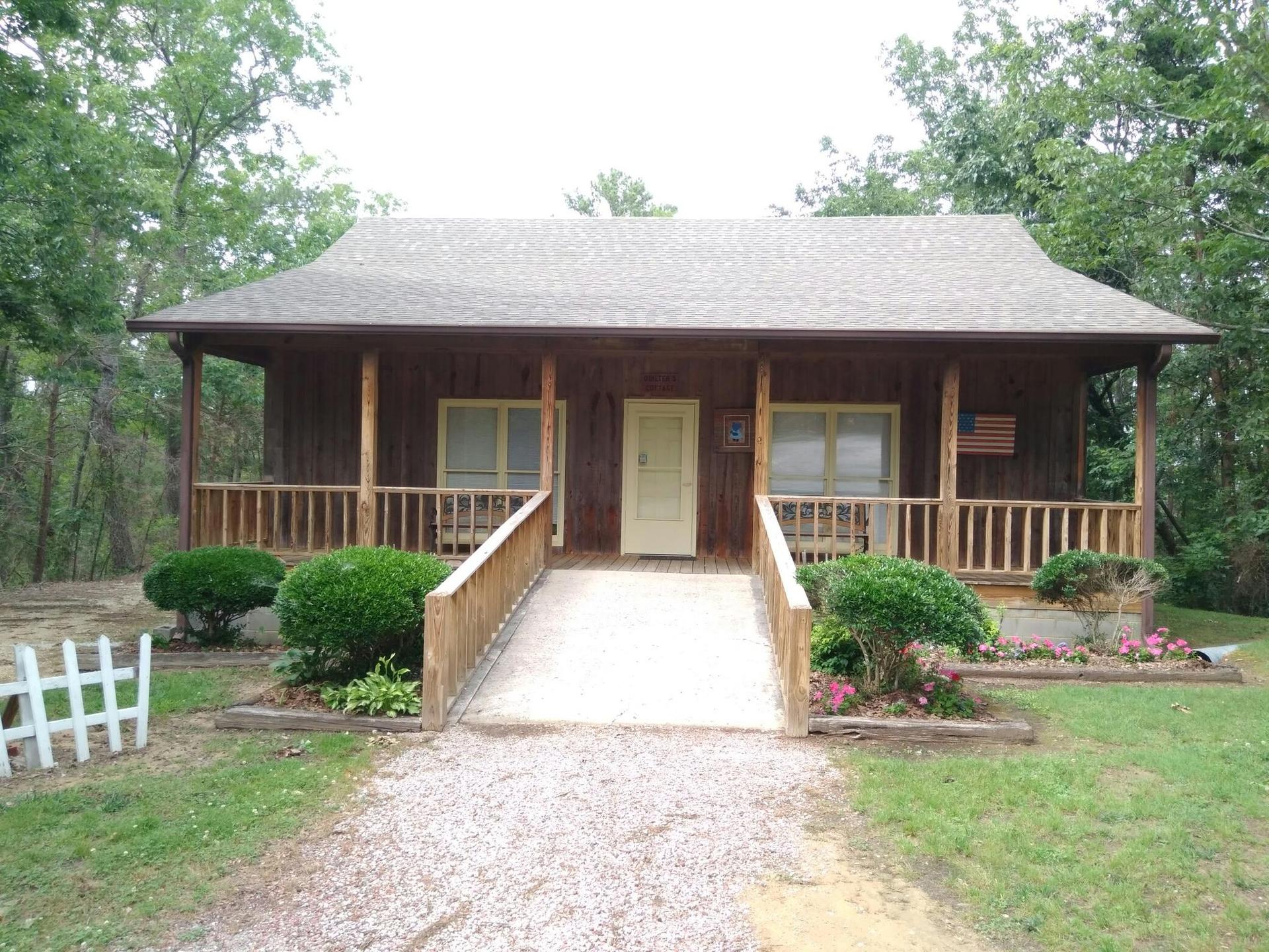 A rustic wooden cabin with a porch and ramp, surrounded by green trees.