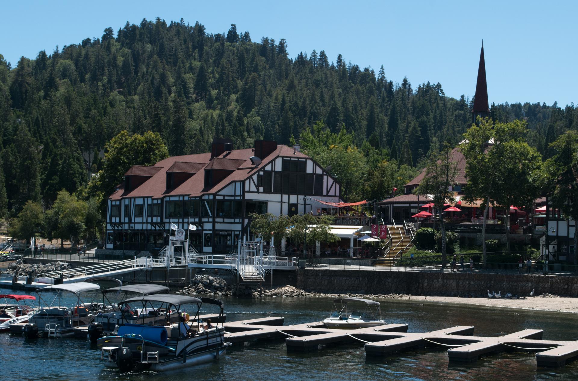 Lakeside buildings with boats docked, surrounded by green trees on a sunny day.