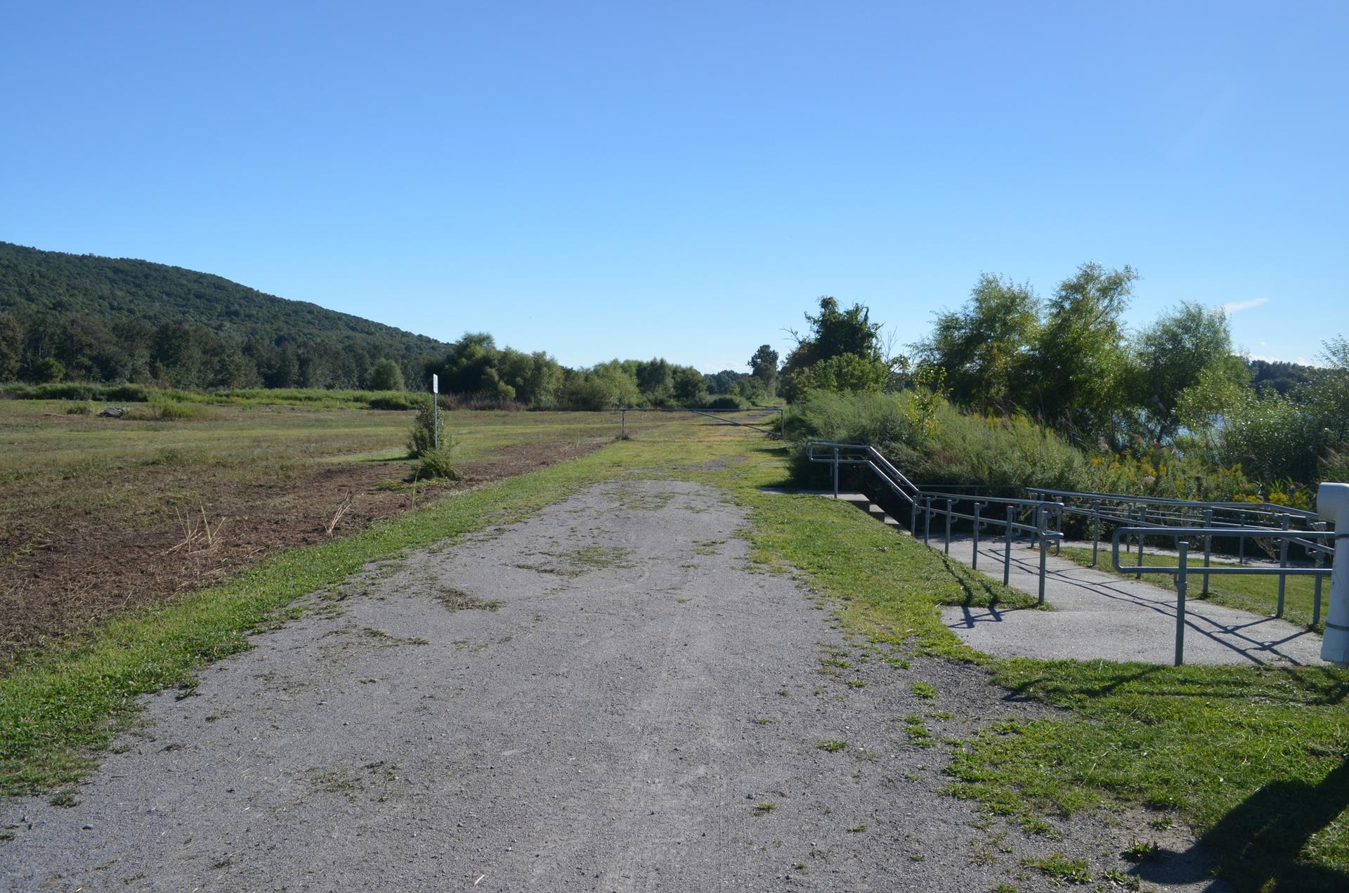 Sunny gravel path, grassy fields, distant mountain, and a ramp with railings.