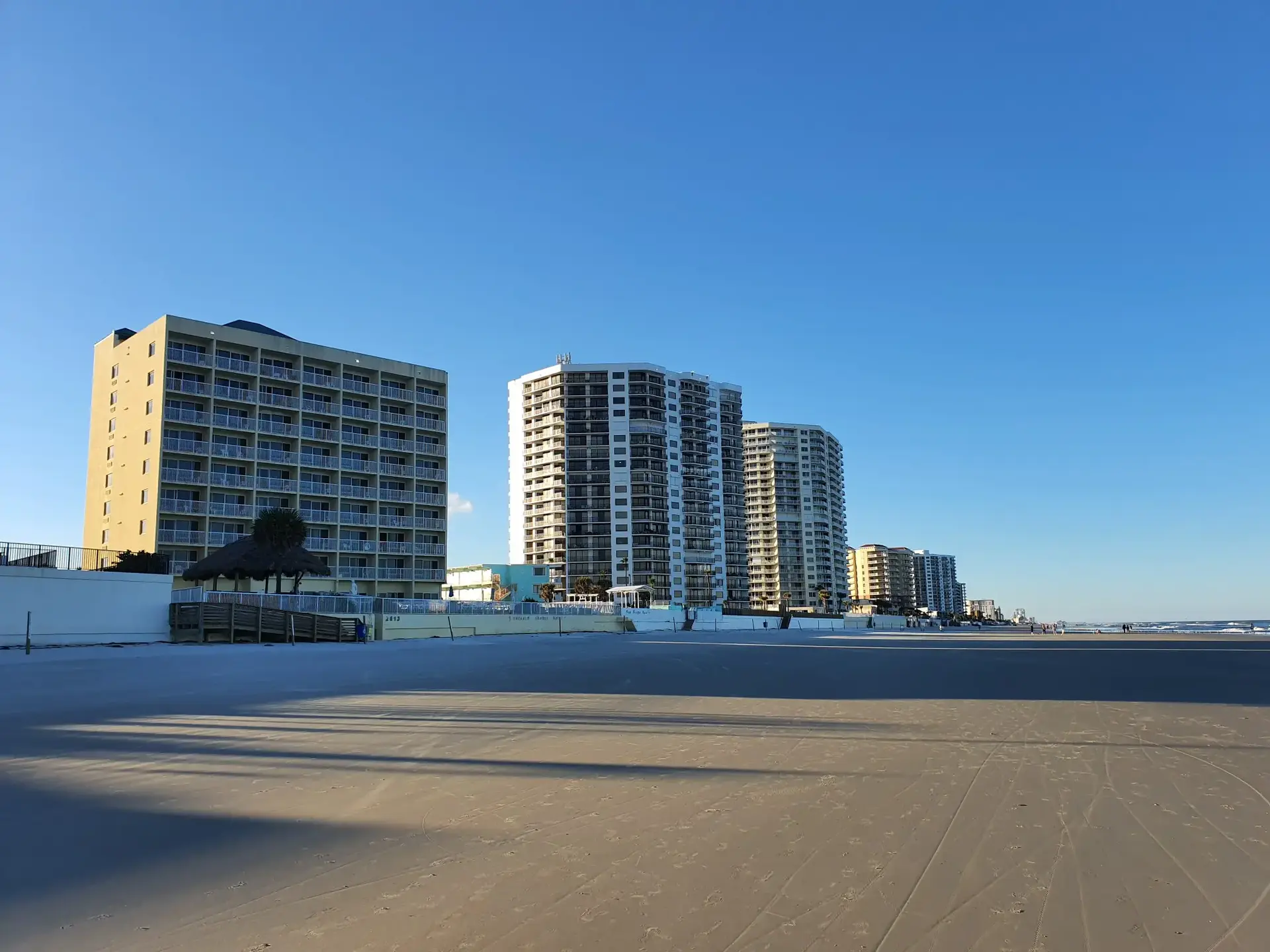 Several tall buildings line a wide, empty beach under a clear blue sky.