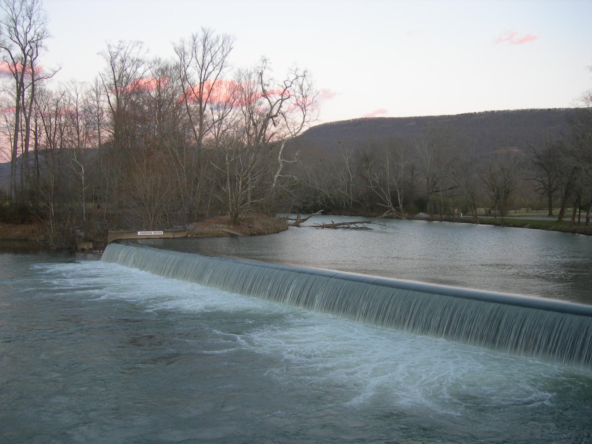 Water flows over a wide dam at dusk, surrounded by bare trees and hills.
