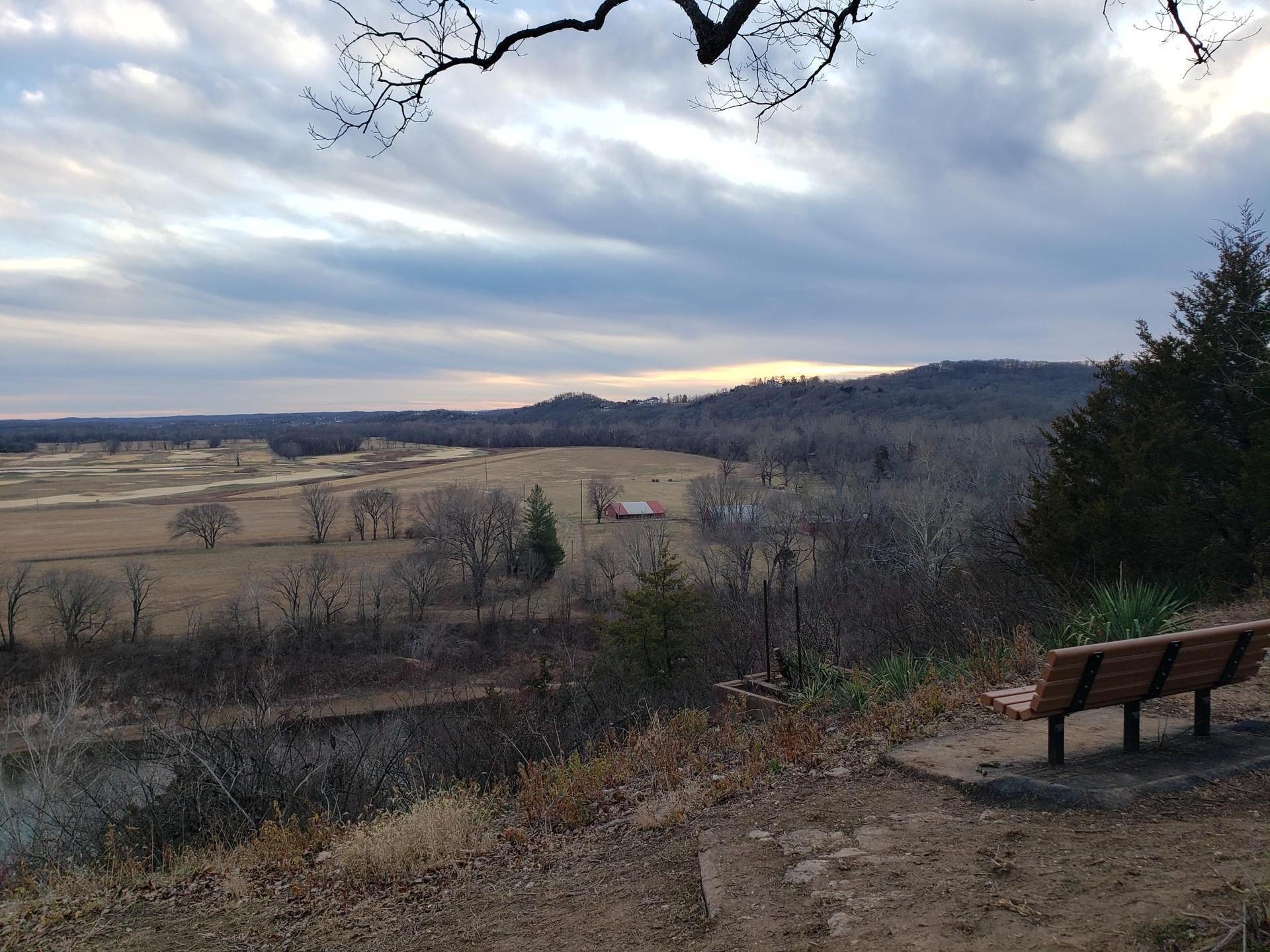 Overlook with a wooden bench facing a valley, bare trees, and cloudy sky.
