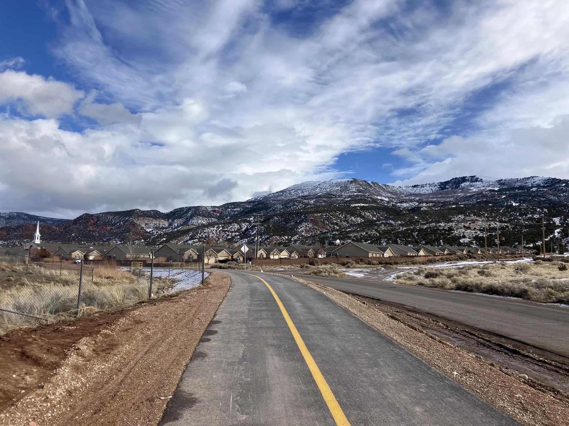 Paved path and road leading towards snow-dusted mountains and houses.