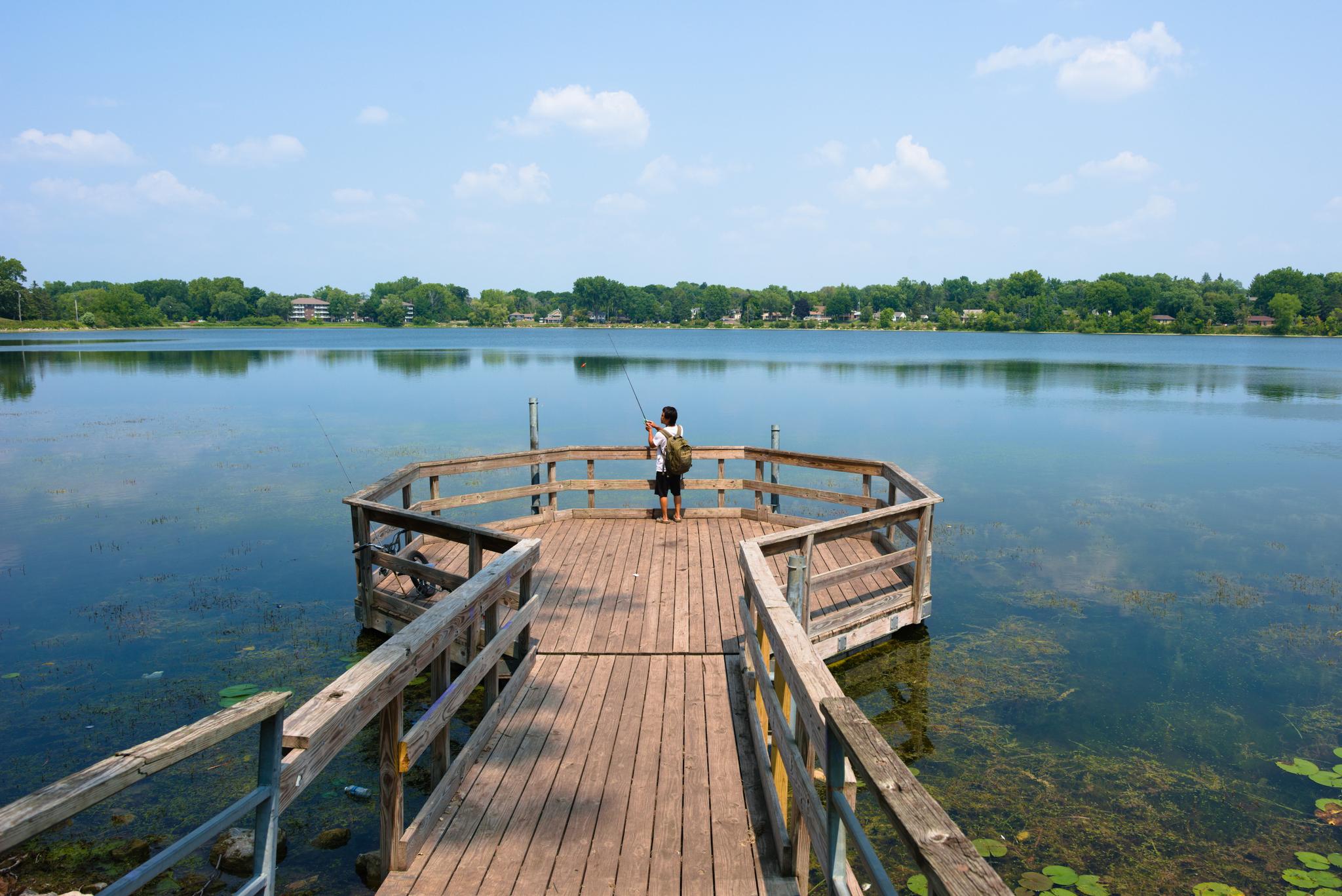 Person fishing from a wooden pier on a calm lake, sunny day.
